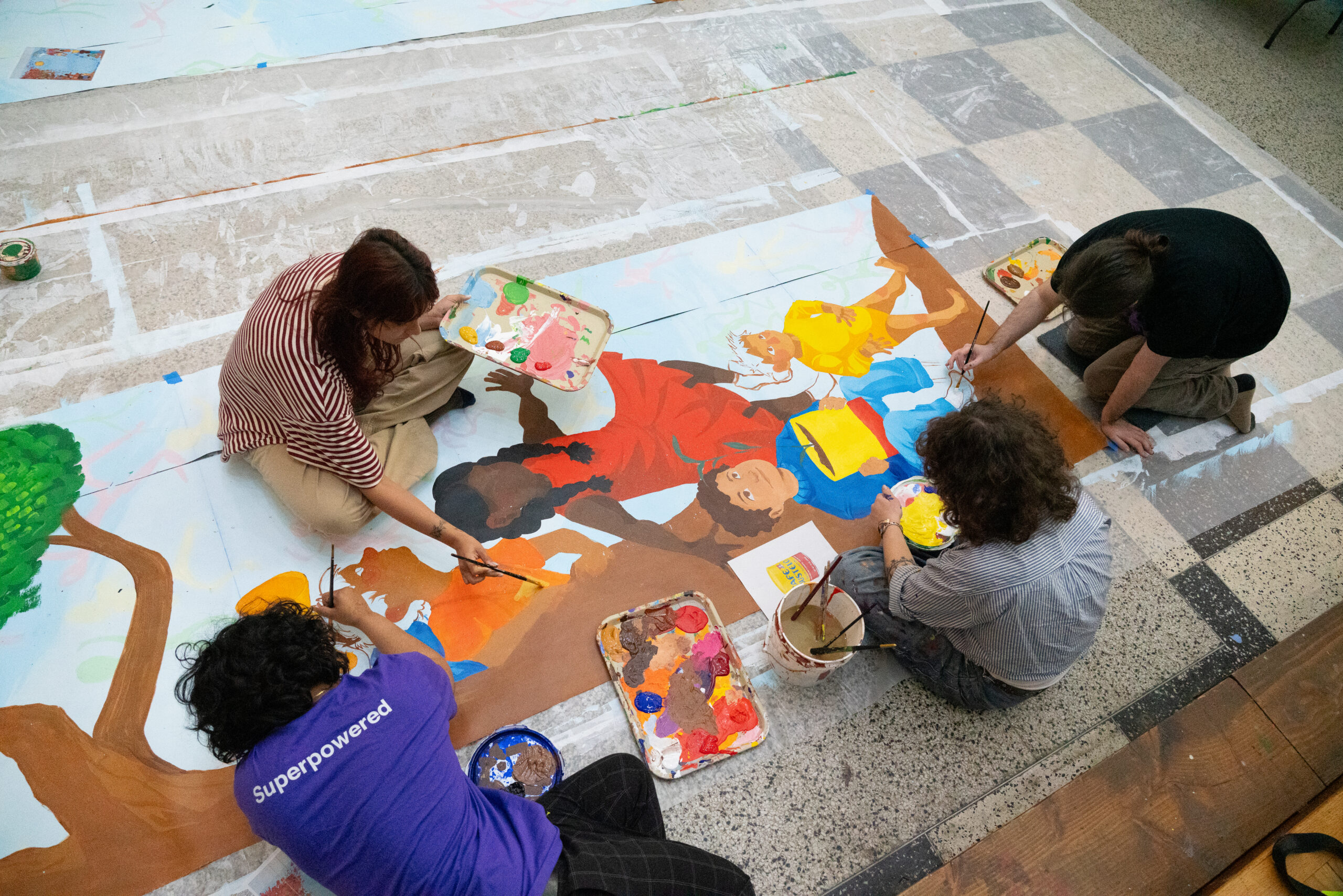 Four people are seated on the floor, engaged in painting a large mural. They are surrounded by various containers of paint and brushes. One person, in a striped shirt, holds a palette, while others are focused on applying color to different sections of the mural, which features figures and vibrant colors. The setting appears to be indoors with a tiled floor.