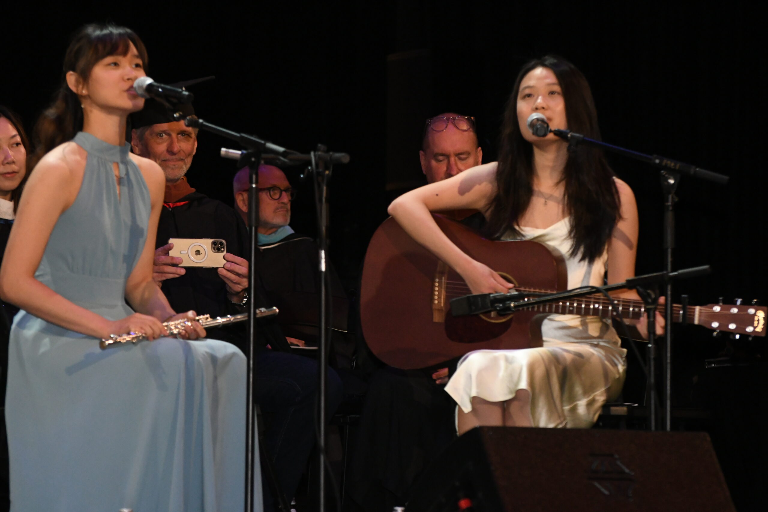 Two female musicians perform on stage. The musician on the left plays the flute and sings, wearing a light blue dress. The musician on the right plays an acoustic guitar, dressed in a white gown. They are accompanied by seated audience members, some wearing academic robes. The setting suggests a performance or ceremony.