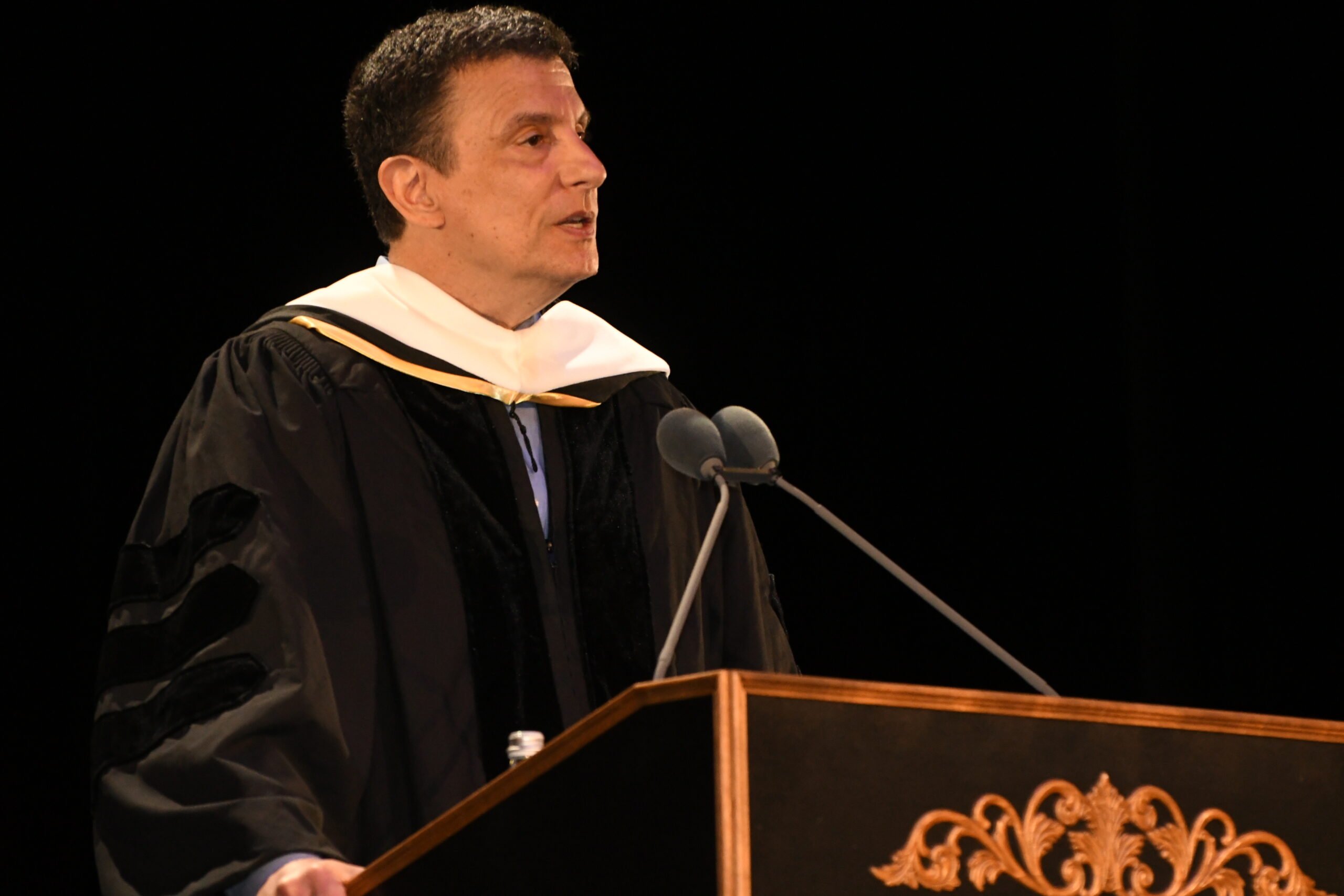 A person in academic regalia speaks at a podium, with a microphone in front. The setting is dark, highlighting the speaker's expression and attire. The podium has ornate detailing and a water bottle is visible on its surface. The speaker appears focused and engaged, addressing an audience.
