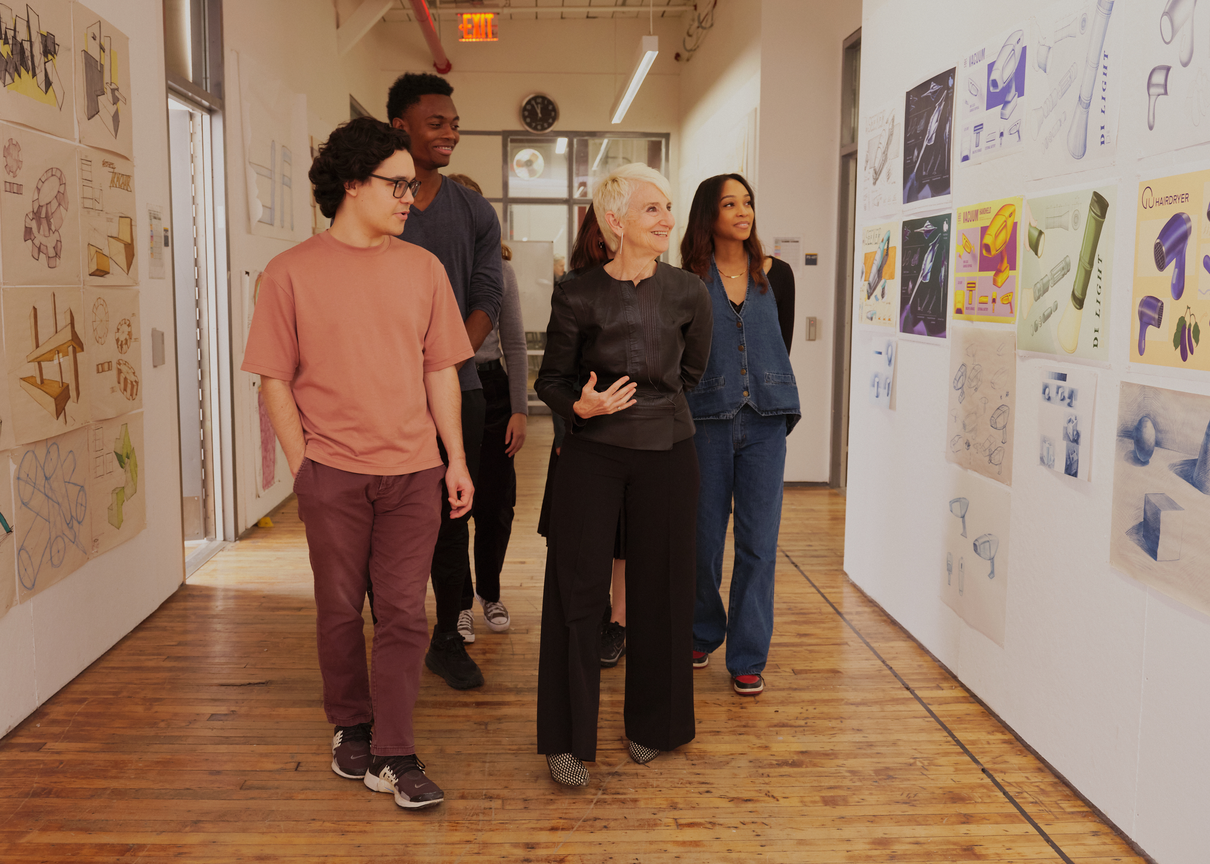 With President Frances Bronet leading, a group of students walks through a brightly lit hallway lined with artwork pinned to white walls.