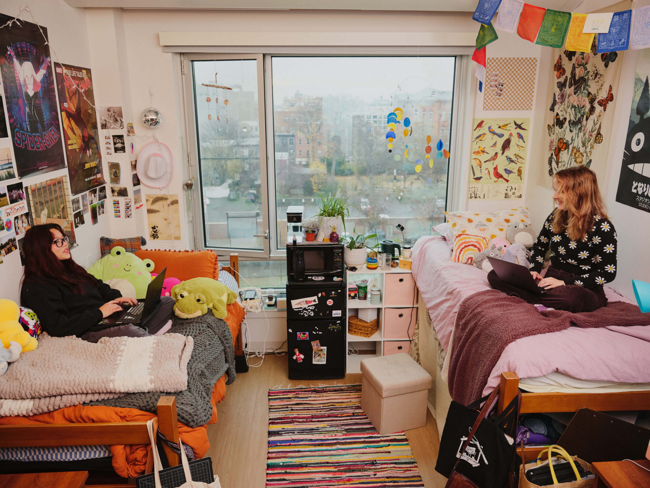 A cozy shared bedroom features two distinct sleeping areas. On the left, a person with long dark hair sits on a bed surrounded by colorful stuffed animals, working on a laptop. On the right, another person with wavy blond hair smiles while using a laptop amidst pillows and a blanket. The room is decorated with posters, photos, and colorful decor, and large windows show a foggy city view outside.