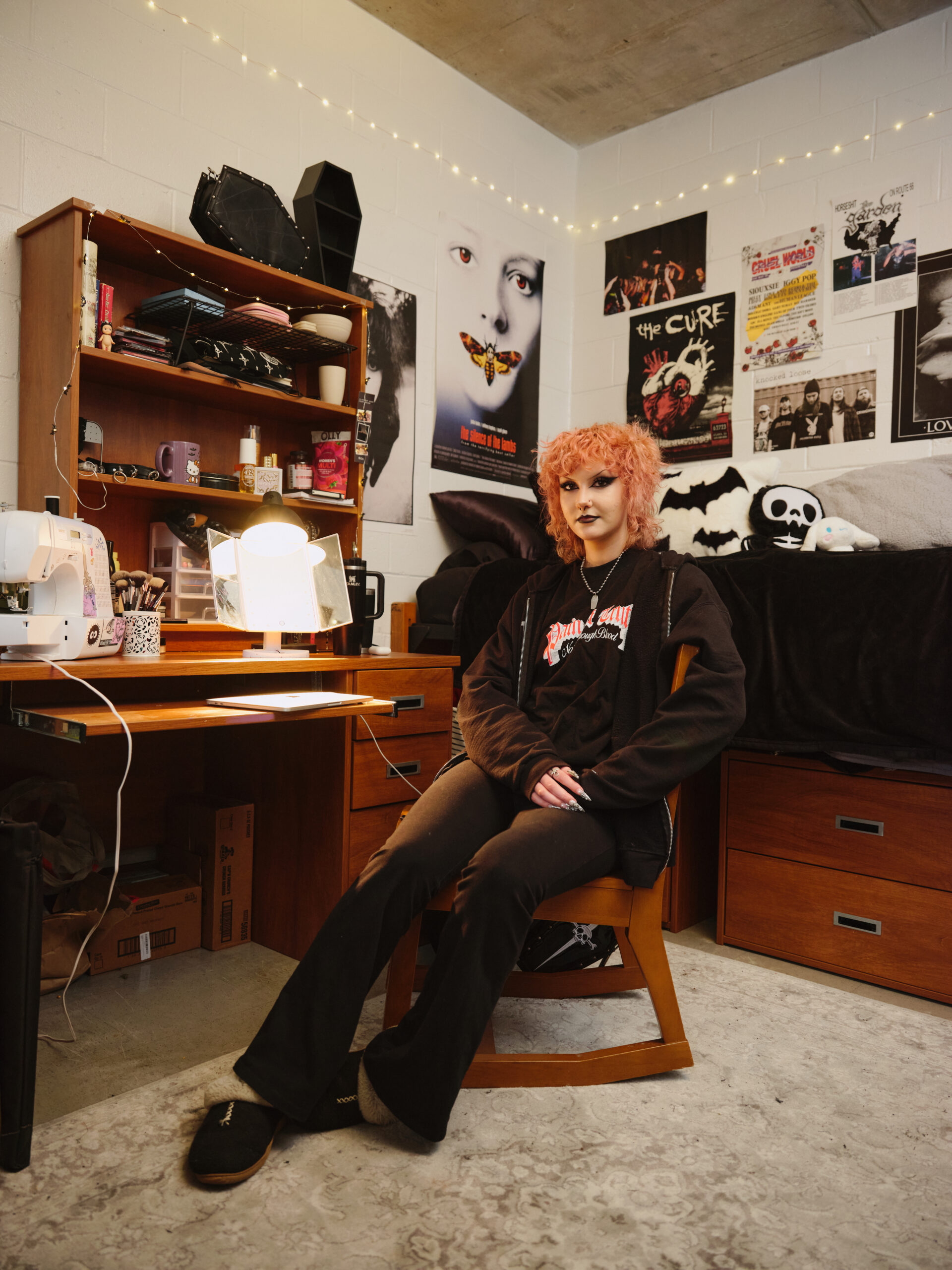 A person with pink, curly hair sits in a wooden chair in a cozy room. The room features a desk with a lamp, a sewing machine, and various decorative items. Posters adorn the walls, including music-related images. A bed with black bedding is in the background, and soft lighting adds a warm atmosphere. The person wears a black hoodie and is resting their hands on their knees.