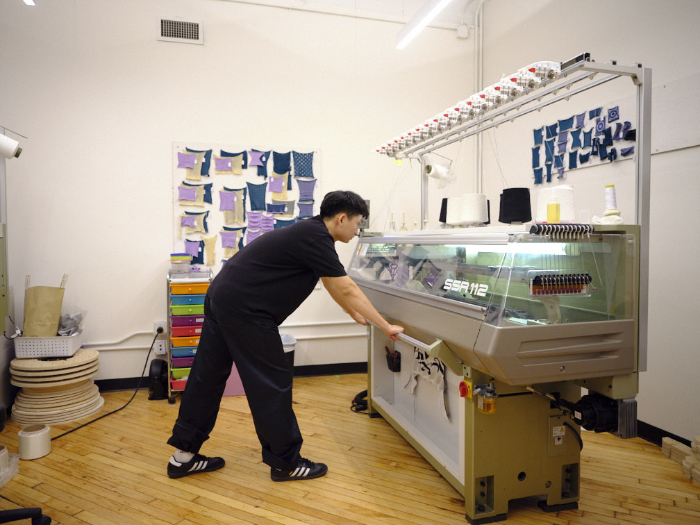 A person is working at a sewing machine in a bright, well-organized studio. The machine features a transparent casing and is labeled "SSR 112." Behind the worker, there is a wall displaying various fabric swatches in different colors, and a shelf with neatly arranged craft supplies. The floor is made of wooden planks, and the overall atmosphere is creative and industrious.