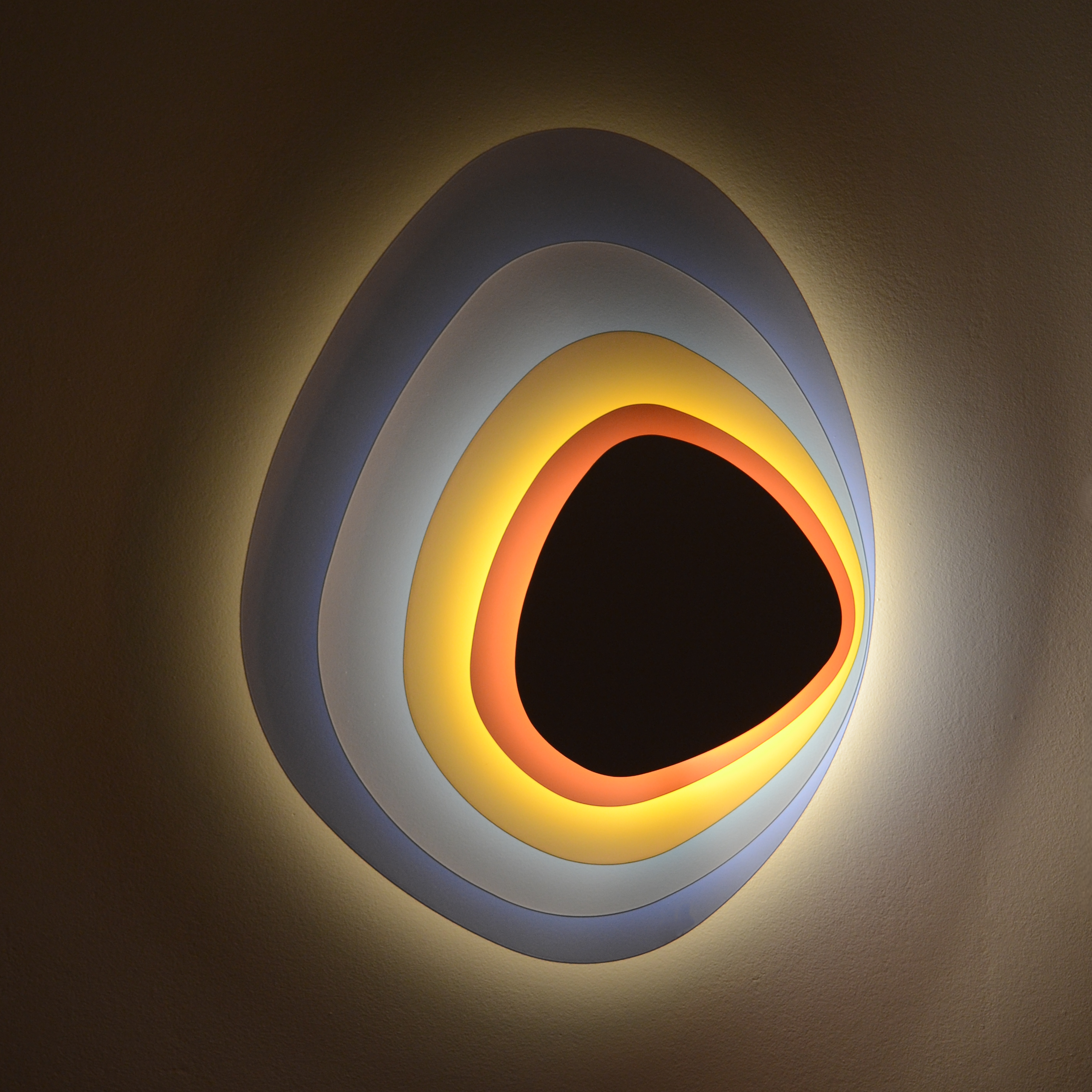 A glowing wall sconce with a rounded organic form made of layers of material. Each layer, from closest to the wall to the most projected, gets gradually smaller as the layers shift from dark blue to pale blue to yellow to orange and finally to black.