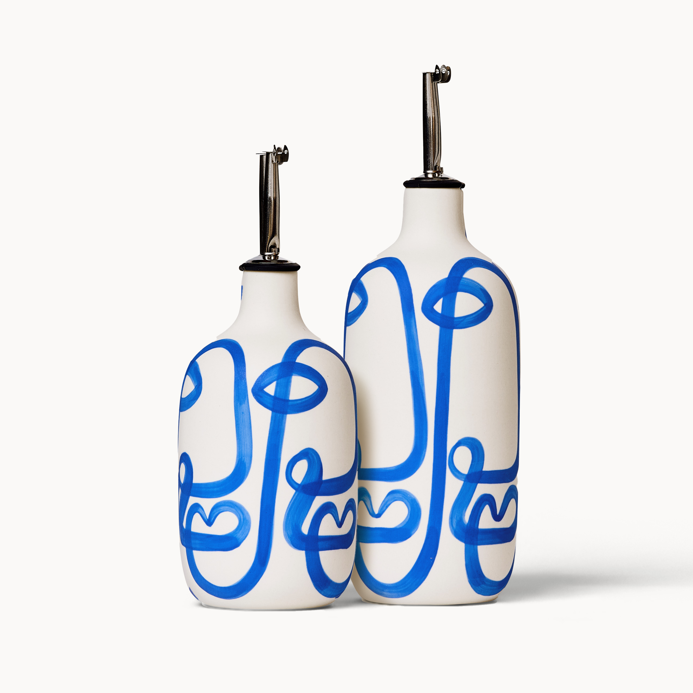 White olive oil cruets. One is long and tall while the other has a shorter and more squat form. They are decorated with gestural blue lines that form an abstract version of a human face.