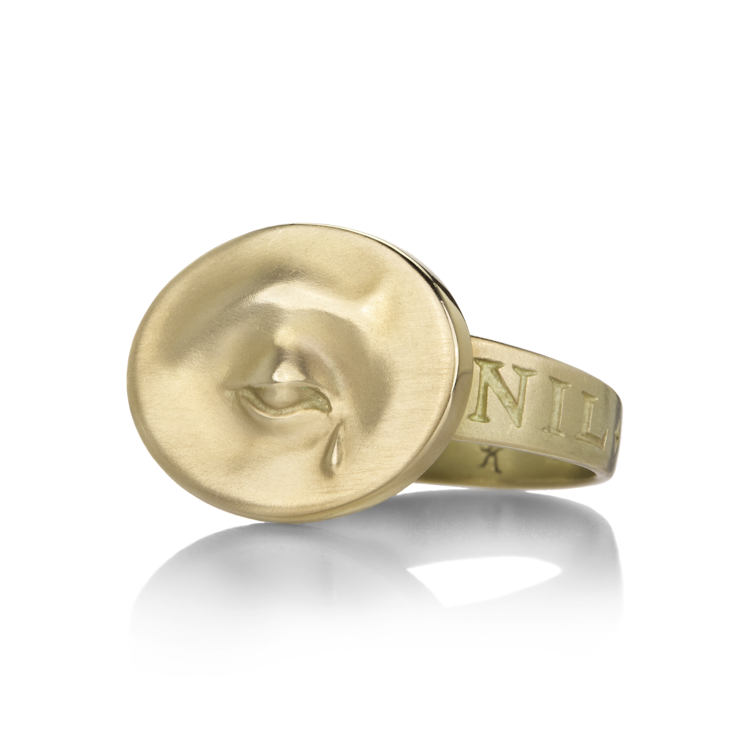 A gold ring. At the center of the ring is a small medallion with a three-dimensional image of a single eye shedding a tear. On the ring's band can be seen a partial inscription reading "NIL."