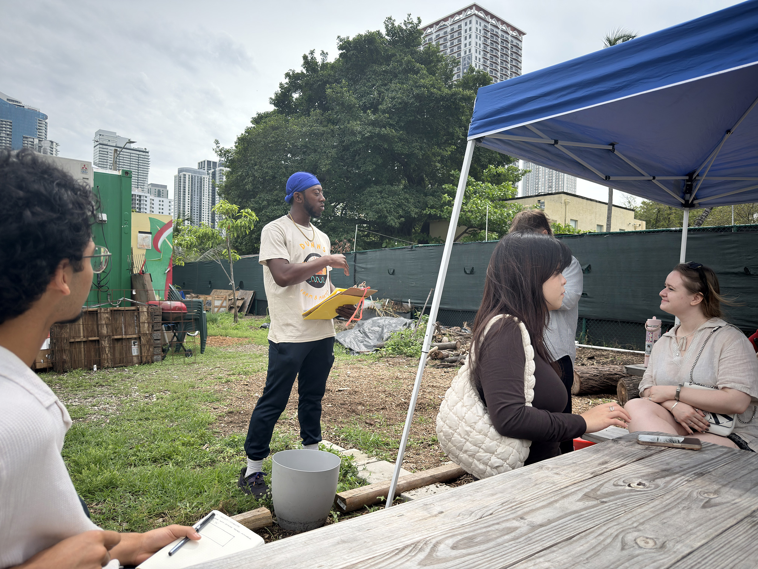 A group of people is gathered outdoors in a community garden. One person, wearing a t-shirt and a blue headband, stands and speaks while holding papers. In the foreground, two people are seated at a wooden table with notebooks. One person observes the speaker, while another engages with someone beside them under a blue tent. The background features urban buildings and greenery, indicating a lively setting.