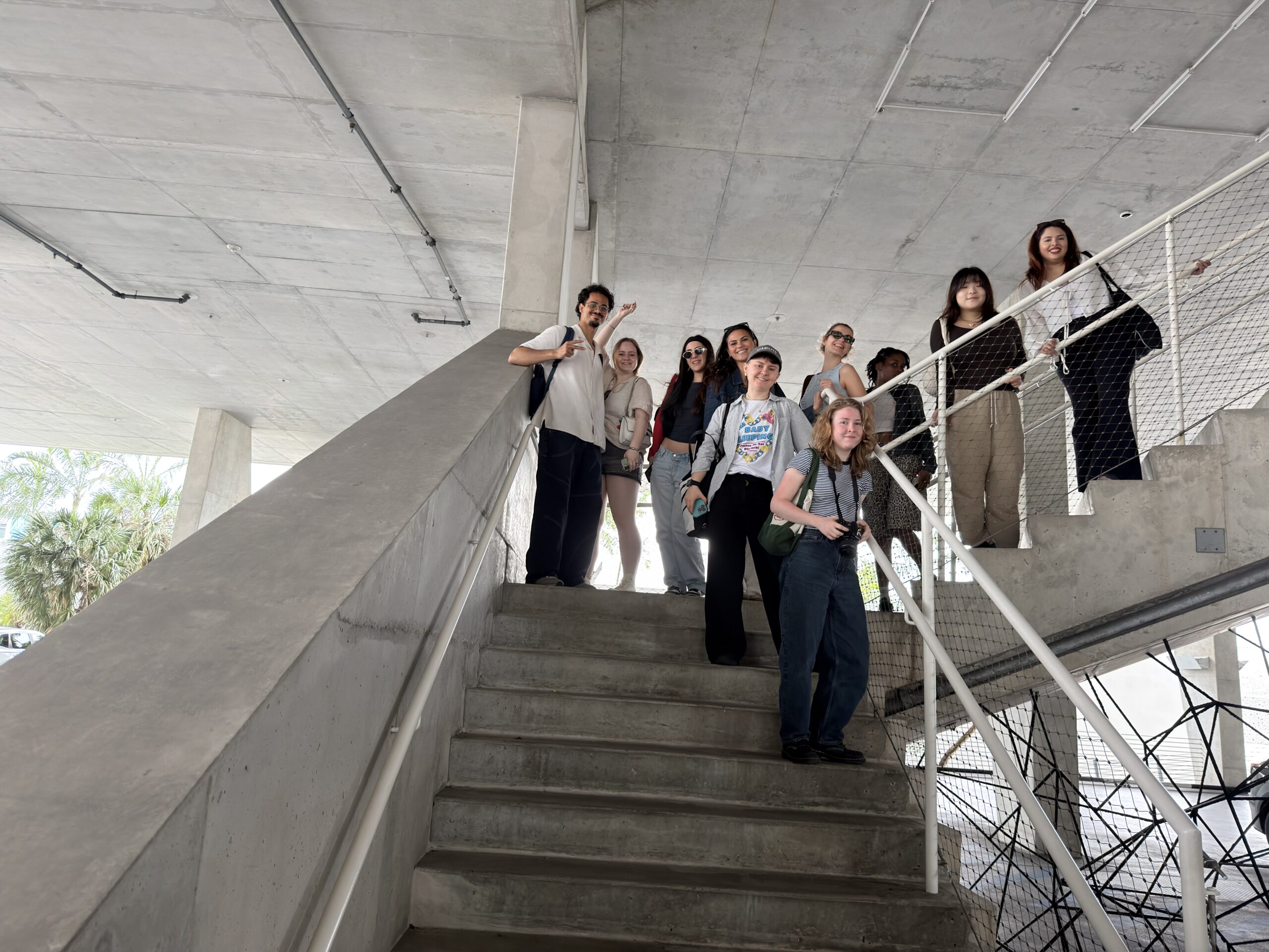 A group of diverse individuals stands on a staircase in a modern building. Some are posing for the camera, smiling and waving. The staircase has concrete steps and a white railing. In the background, there are palm trees visible through the open space at the bottom of the staircase. The environment is bright and airy, indicative of a contemporary architectural style.
