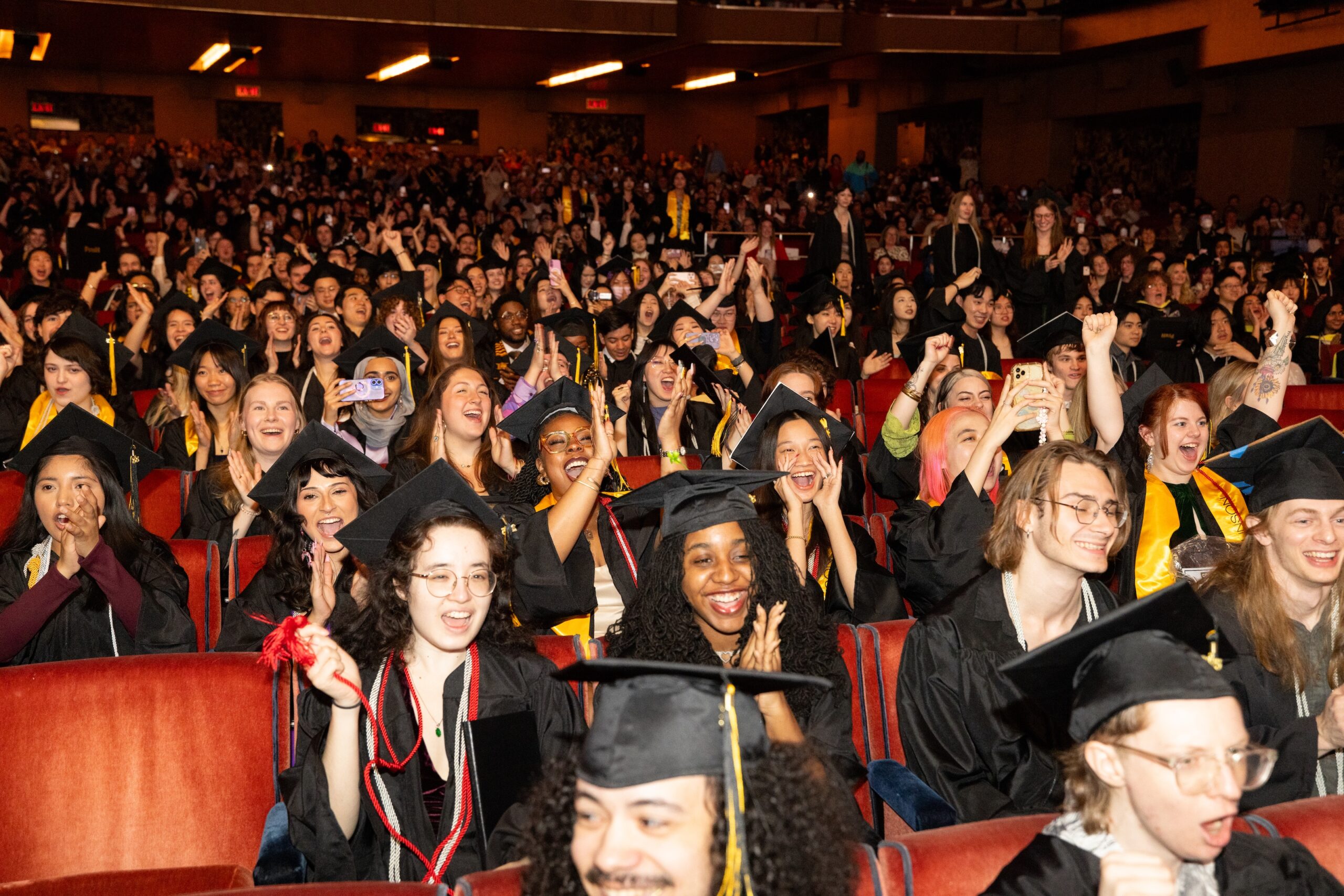 A large audience of graduates in black caps and gowns is cheering and celebrating in a theater. Many are clapping, smiling, and shouting with excitement. The atmosphere is joyful and vibrant, with some individuals holding phones to capture the moment. The audience features a diverse group of people, with a backdrop of red theater seats and dim lighting.