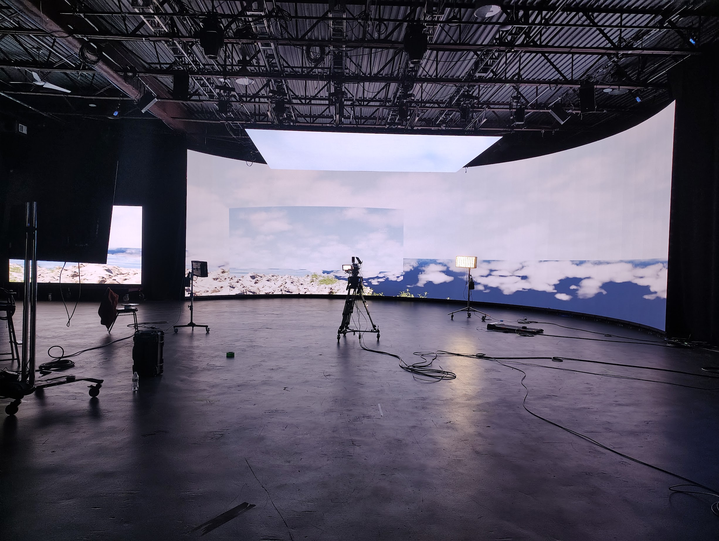 A spacious studio with a curved LED backdrop displaying a scenic landscape with clouds and water. In the foreground, there are various pieces of equipment, including a camera on a tripod, lighting fixtures, and cables scattered on the black floor. A chair is visible, and a small suitcase is positioned nearby. The overhead structure features exposed beams and lighting elements.