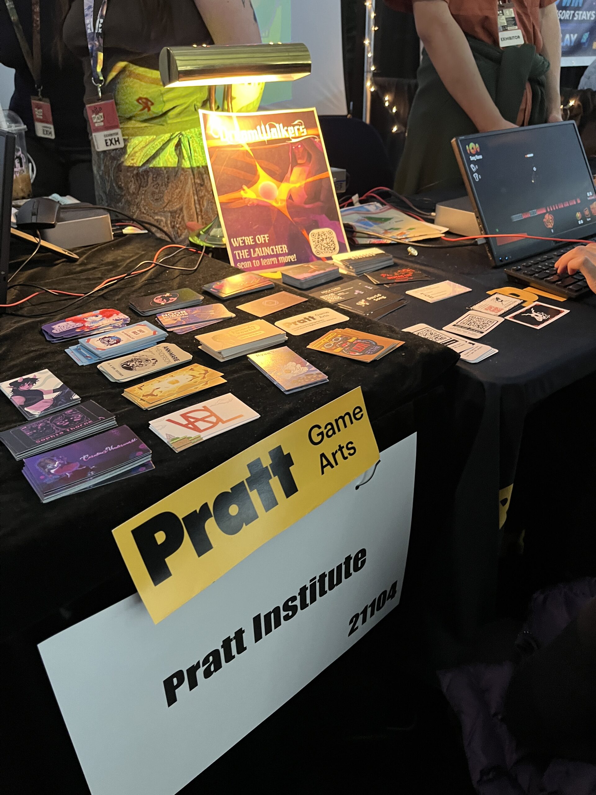 A table is set up with various colorful game cards displayed in front. A bright lamp illuminates a poster for 