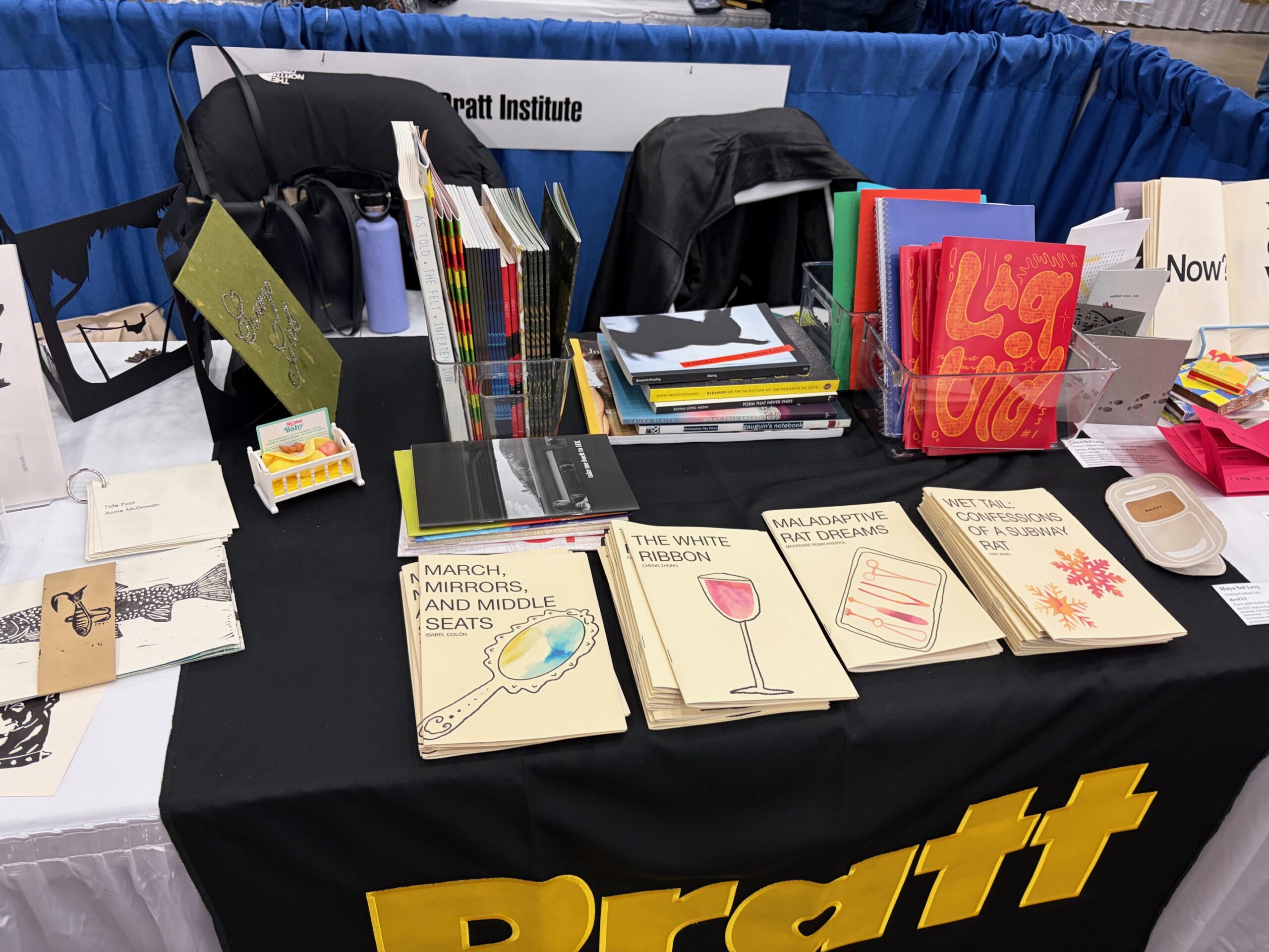 A tabletop display featuring various books and notebooks from Pratt Institute. The table is covered with a black cloth that has "Pratt" written in yellow. There are several stacks of books, some open and some closed, along with colorful notebooks in a clear container. A small display of items resembling eggs is visible, alongside other unique publications. A backpack and a water bottle are partially seen in the background.