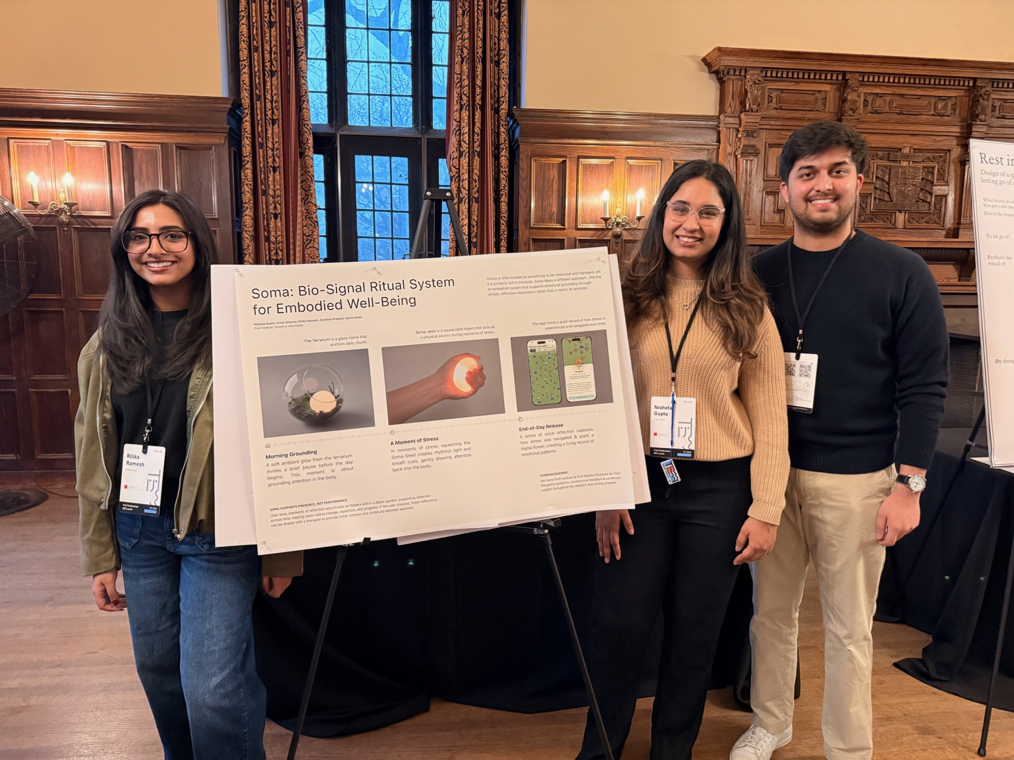 Three individuals are standing in front of a display board titled "Soma: Bio-Signal Ritual System for Embodied Well-Being." One person is on the left, wearing glasses and a green sweater, while the middle person is wearing a beige sweater and the person on the right is in a black sweater. The display board features text and images related to the project, which seems to focus on well-being rituals. The background includes wooden paneling and large windows.