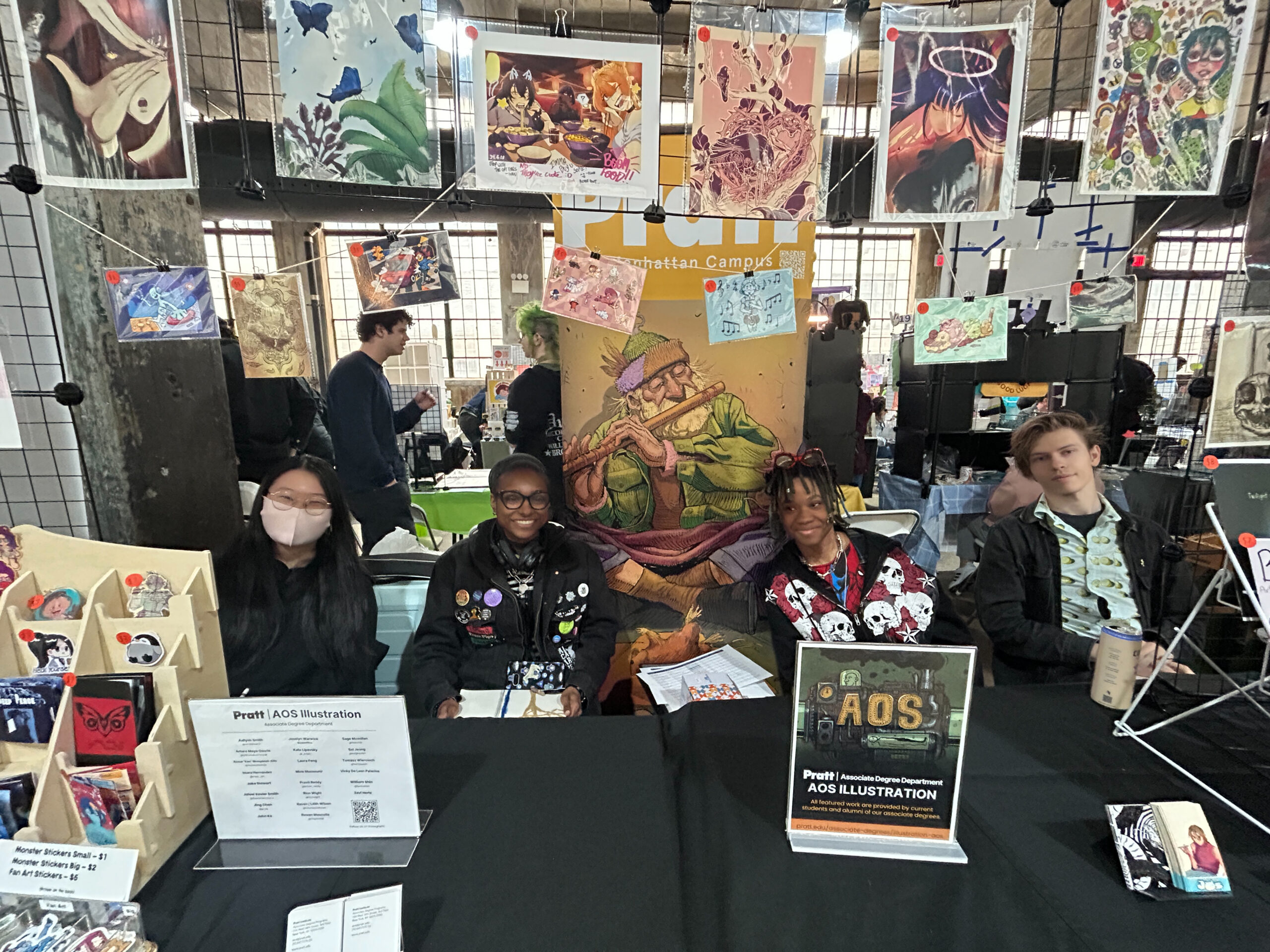 A group of four artists is seated at a table in a busy convention space, surrounded by various art prints displayed above. They are showcasing their work for Pratt's AOS Illustration program. Two of the artists wear masks, while another has colorful braids and is dressed in a patterned shirt. The table features a small display of stickers for sale, and a sign indicating their association with Pratt Institute.