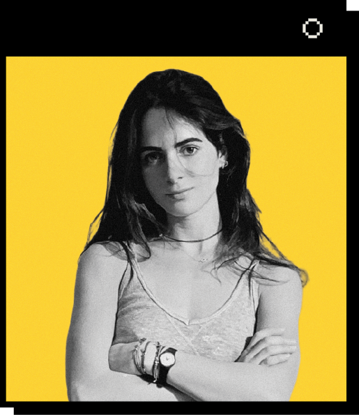 Portrait of Leda Tsoutreli against a yellow background. Leda has long, dark hair and stands with her arms crossed, wearing a gray tank top and multiple bracelets and a watch.