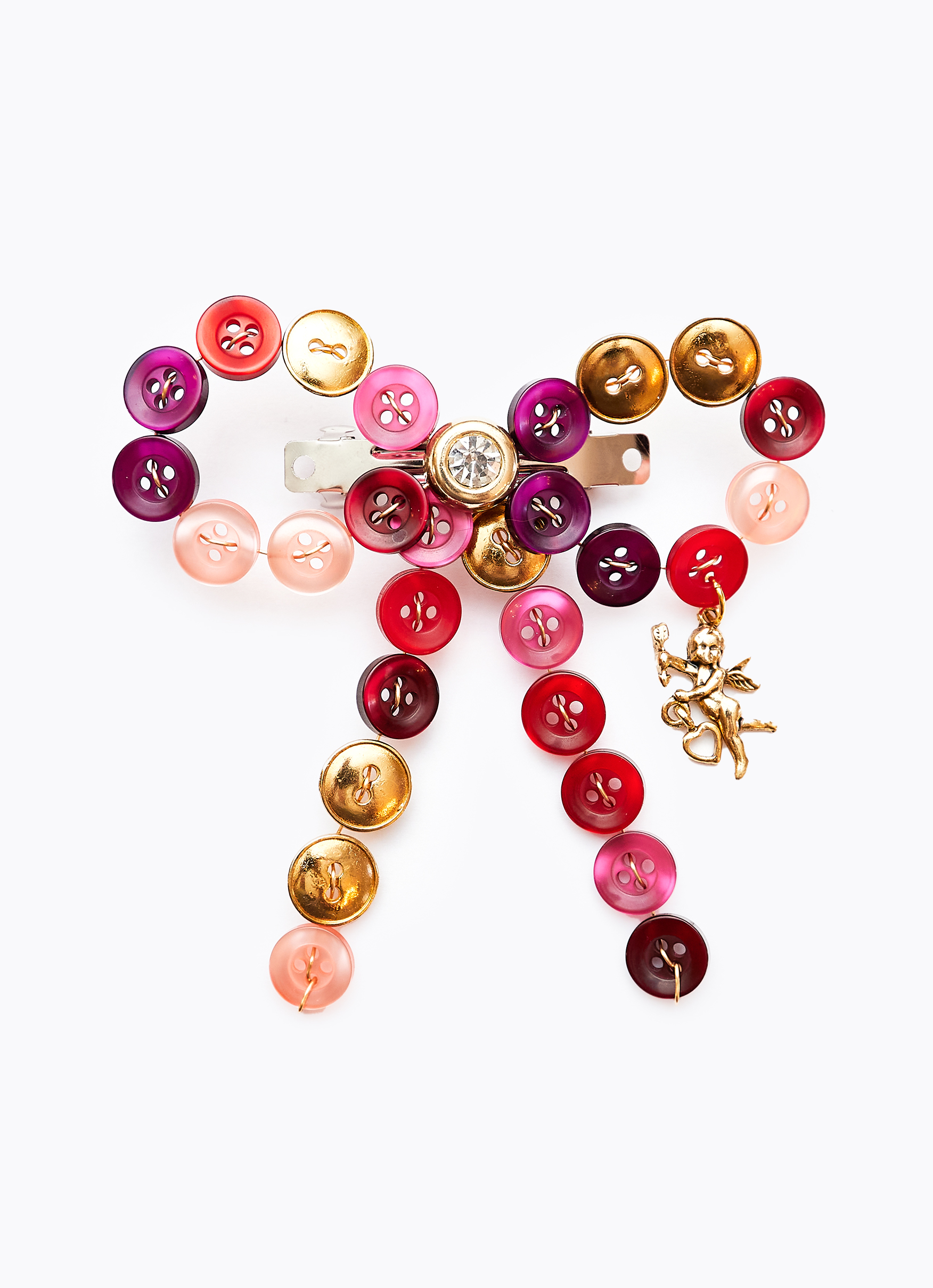 A silver metal hair clip to which is affixed a bow-shaped arrangement of buttons in hues of gold, red, pink, and purple. Dangling from the bow is a golden charm shaped like cupid.