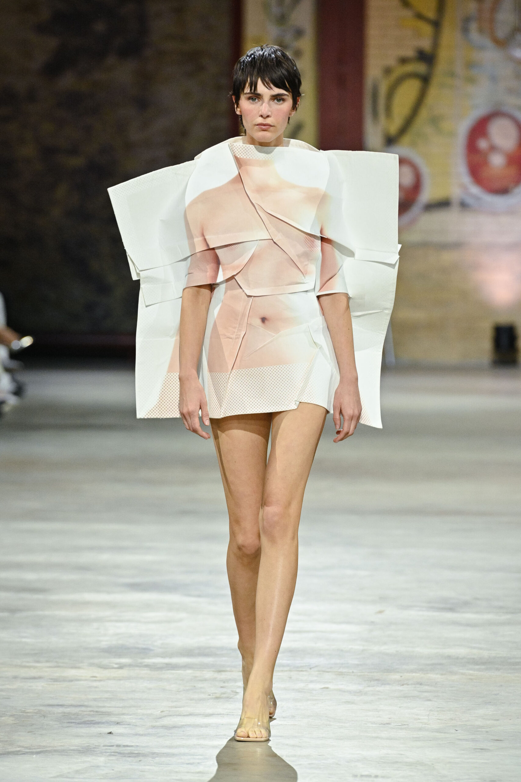 A model walks down a runway wearing a creative and avant-garde outfit that features large, sculptural elements resembling fabric drapes. The outfit has a pale color palette with skin-toned graphics, emphasizing a unique silhouette. The venue has an industrial feel, with a polished concrete floor and a backdrop that includes colorful murals or patterns. The model has short, dark hair and wears minimal accessories, allowing the outfit to be the focal point.