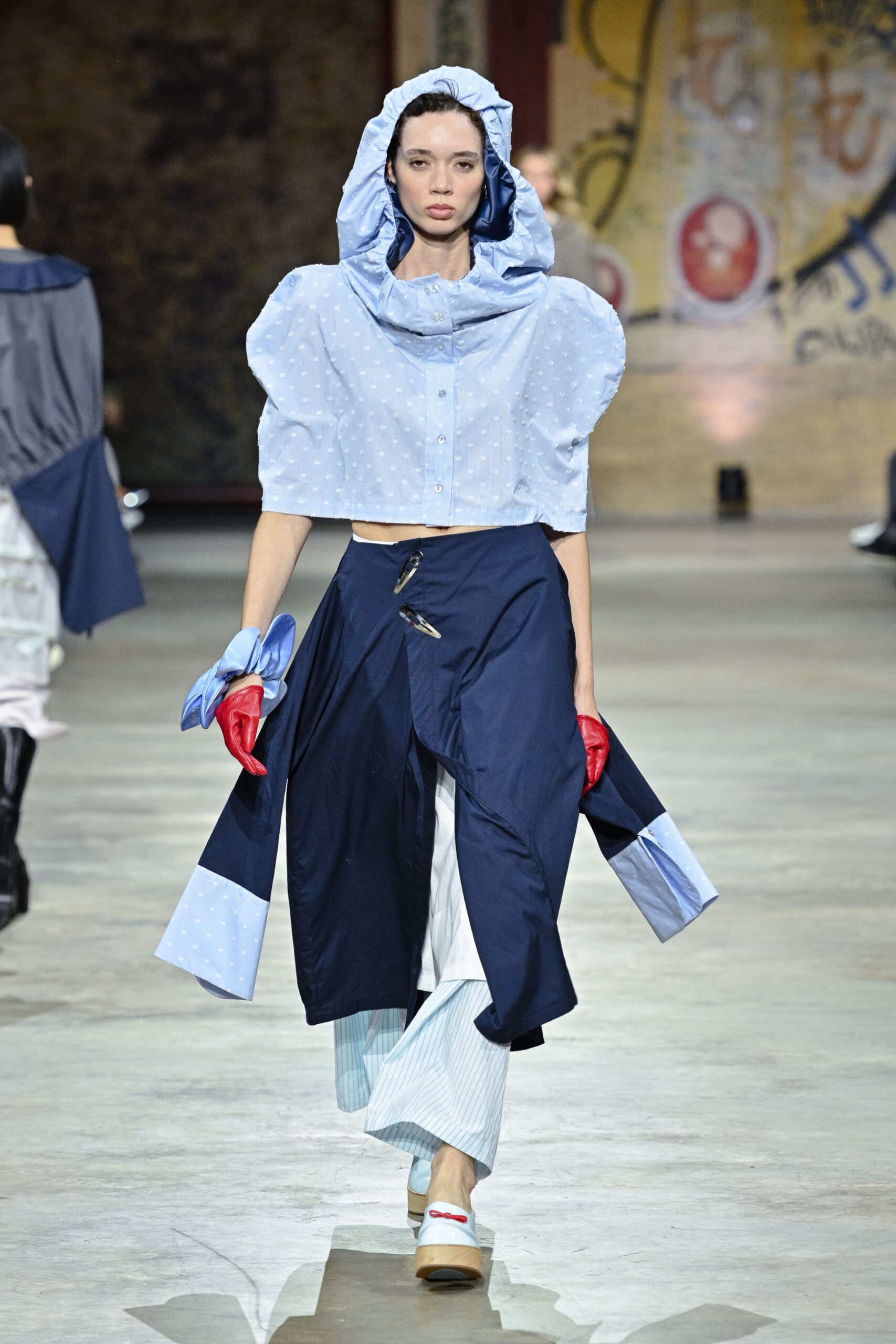 A model walks down a runway wearing a unique outfit featuring a light blue cropped top with a large hood and puffed sleeves, paired with a dark blue skirt that has a wrap design. She accessorizes with red gloves and a blue bow tied around her wrist. The setting has an industrial feel, with a graffiti-style mural in the background.