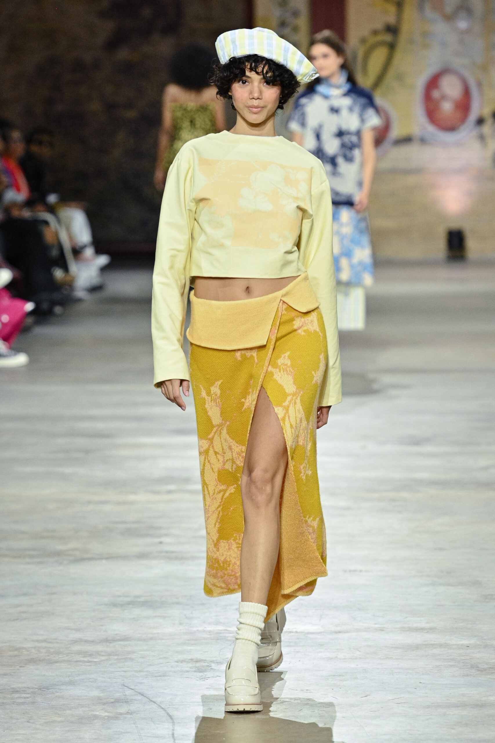 A model walks down a runway wearing a light yellow cropped sweater with floral designs and a long, yellow skirt with a side slit and floral patterns. She accessorizes with a light blue and white checked beret and white platform shoes with ribbed socks. In the background, other models in various outfits can be seen. The setting appears to be an indoor fashion show.