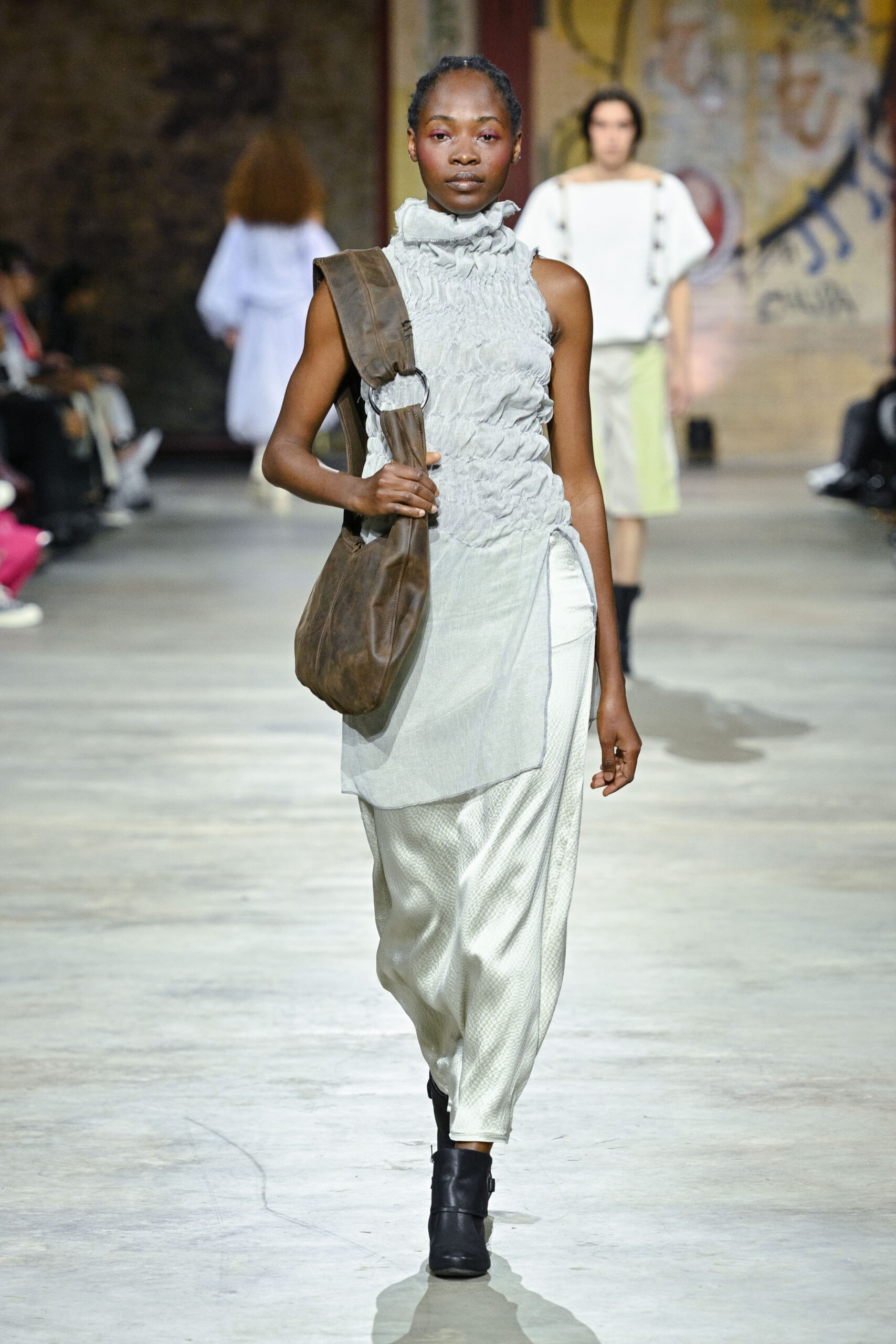 A model walks down a runway wearing a textured, light gray sleeveless top with a high collar, paired with loose, pearl-colored pants. She carries a brown shoulder bag and wears black ankle boots. In the background, other models are visible, and the setting features an industrial, art-inspired backdrop. The audience is seated along the sides of the runway.