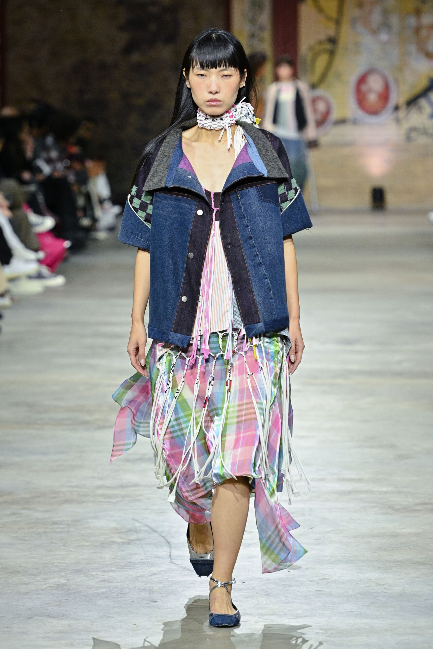 A model walks down the runway wearing a layered outfit featuring a denim jacket with short sleeves and a plaid skirt with colorful fringe. The look is accessorized with a decorative neckpiece. The background shows seated audience members watching the fashion show in a spacious venue.