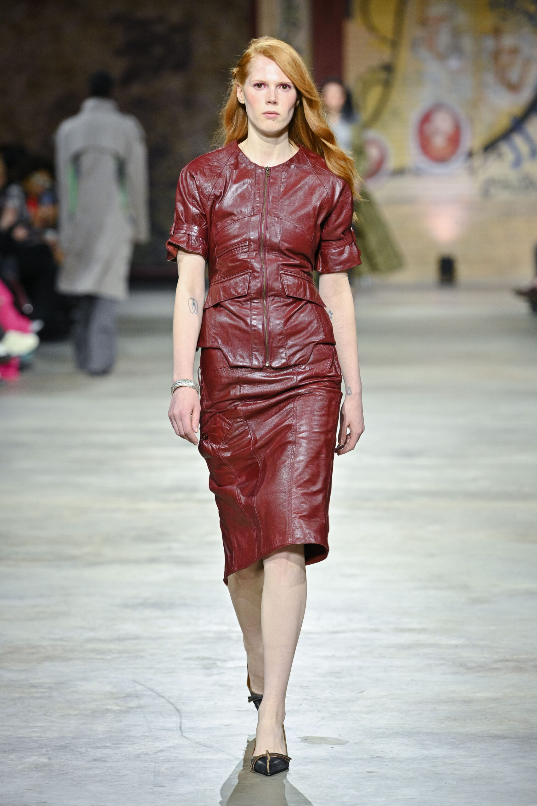 A model walks down a runway wearing a fitted, red leather dress with a zipper at the front and a peplum waist. The dress has short sleeves and a sleek silhouette. The setting is an industrial-looking space with a blurred audience in the background and muted colors behind the model. She has long, wavy red hair and wears black heels.