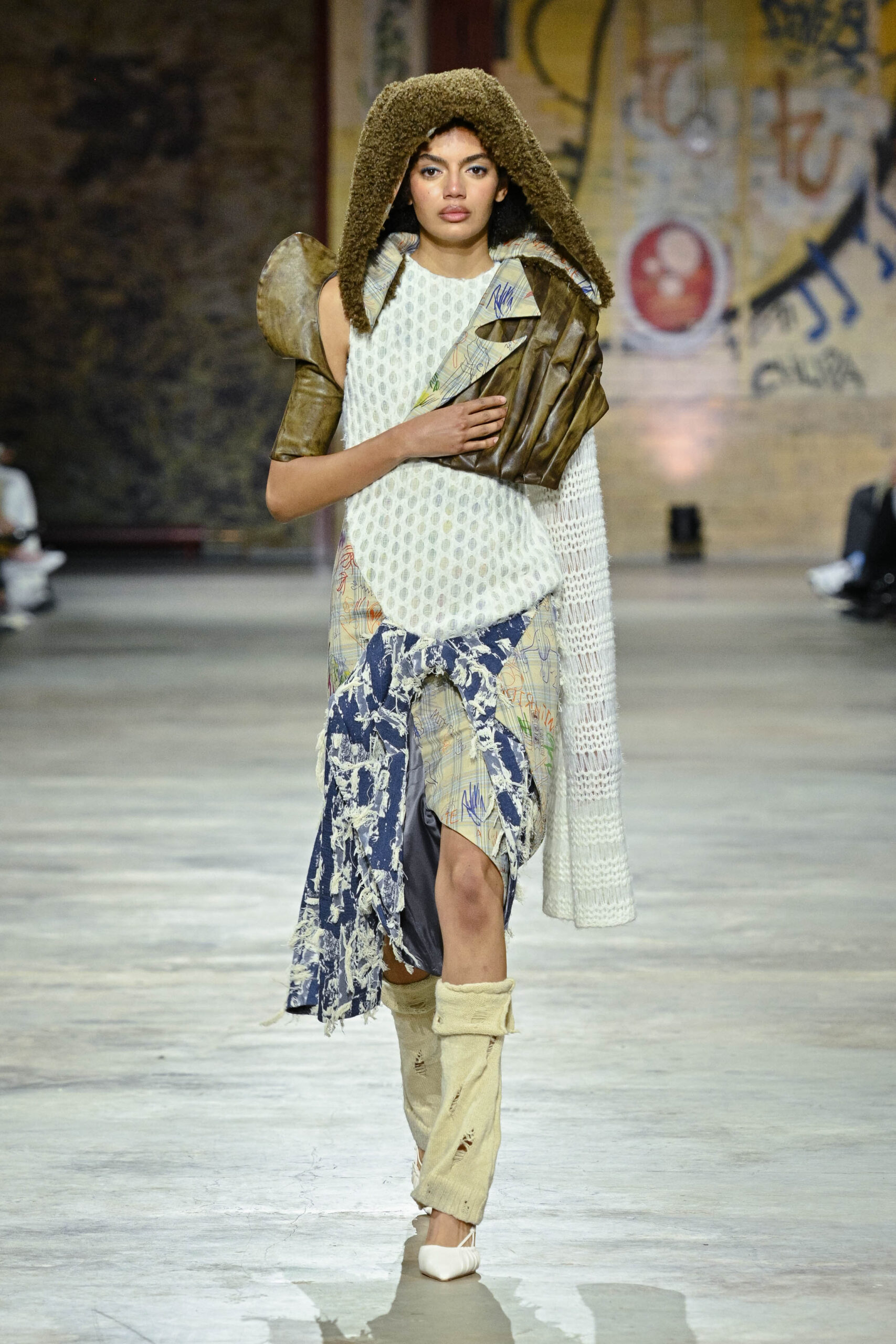A model walks down a runway, wearing a unique outfit featuring a textured white top with a patterned fabric draped over it. The outfit includes a brown shoulder piece, a furry hood, and layered garments with diverse fabrics and patterns. The model's left hand is positioned on her hip while her right hand gently clasps the fabric. The background shows a blurred audience and a graffiti-style wall.
