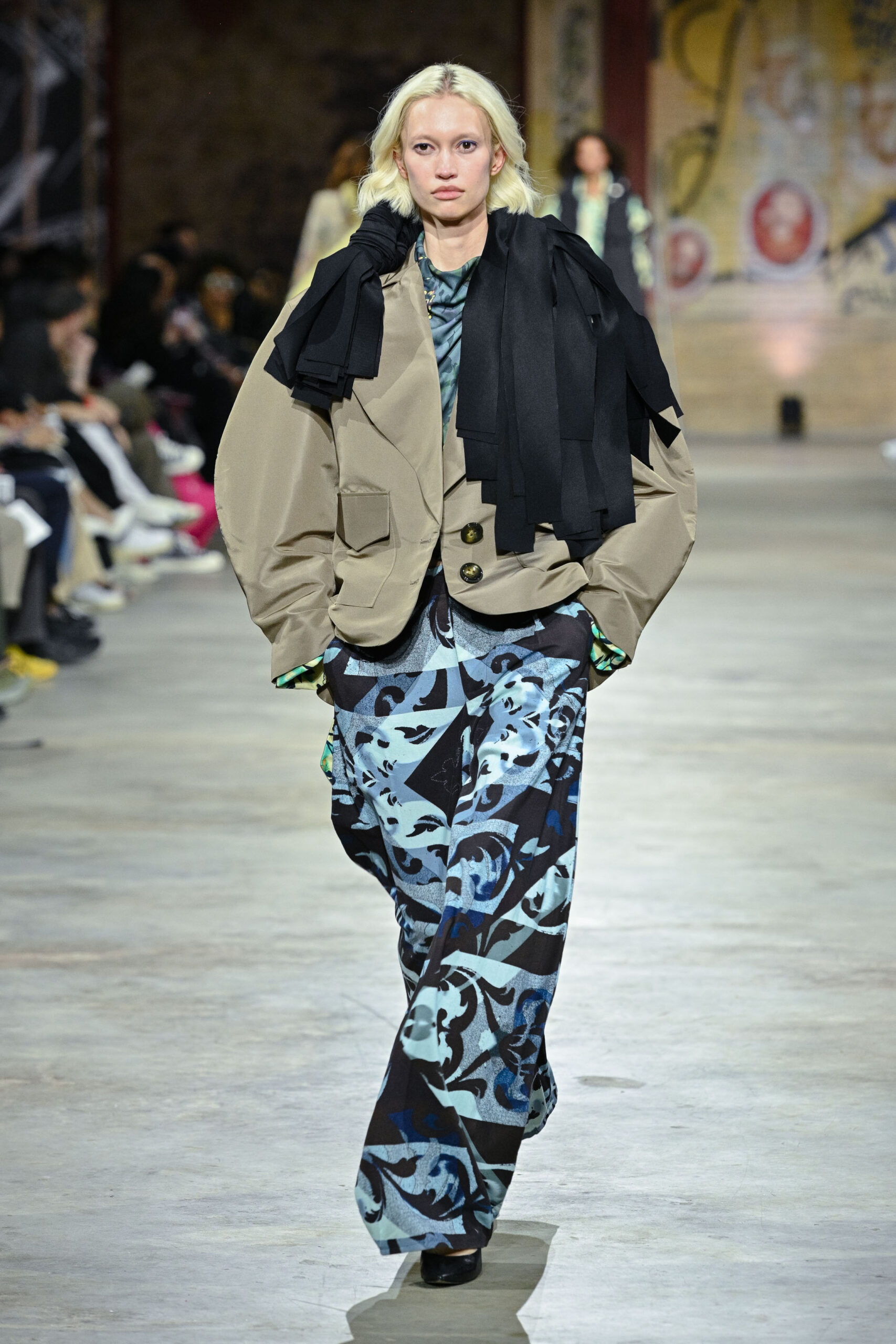 A model walks down a runway, showcasing an oversized beige jacket layered with black fabric draped over one shoulder. She wears a blue and black patterned pant with a hint of vibrant green peeking from underneath. The background is filled with seated spectators, and the setting has an industrial aesthetic with urban artwork on the walls.