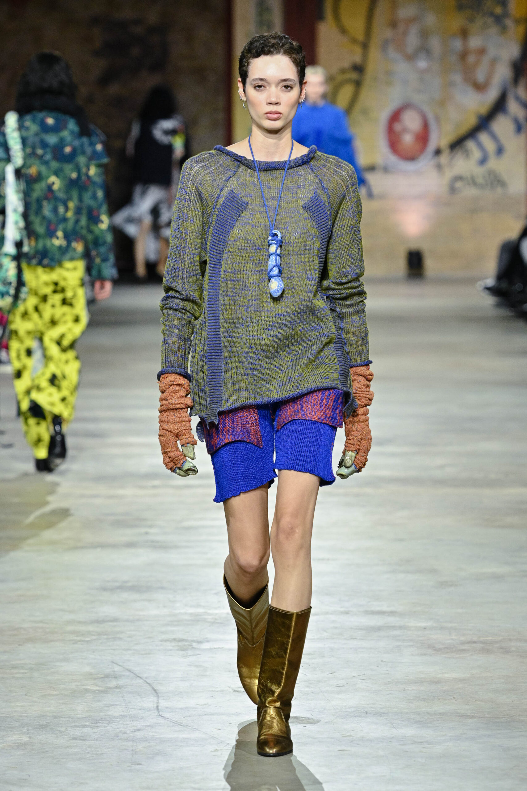 A model walks down a runway wearing a textured olive-green and blue sweater, blue shorts, and brown knitted gloves. She has short, curly hair, and accessorizes with a blue necklace. The background features other models and a colorful, graffiti-style mural on the wall. The runway appears to be in a large, industrial space with a concrete floor.