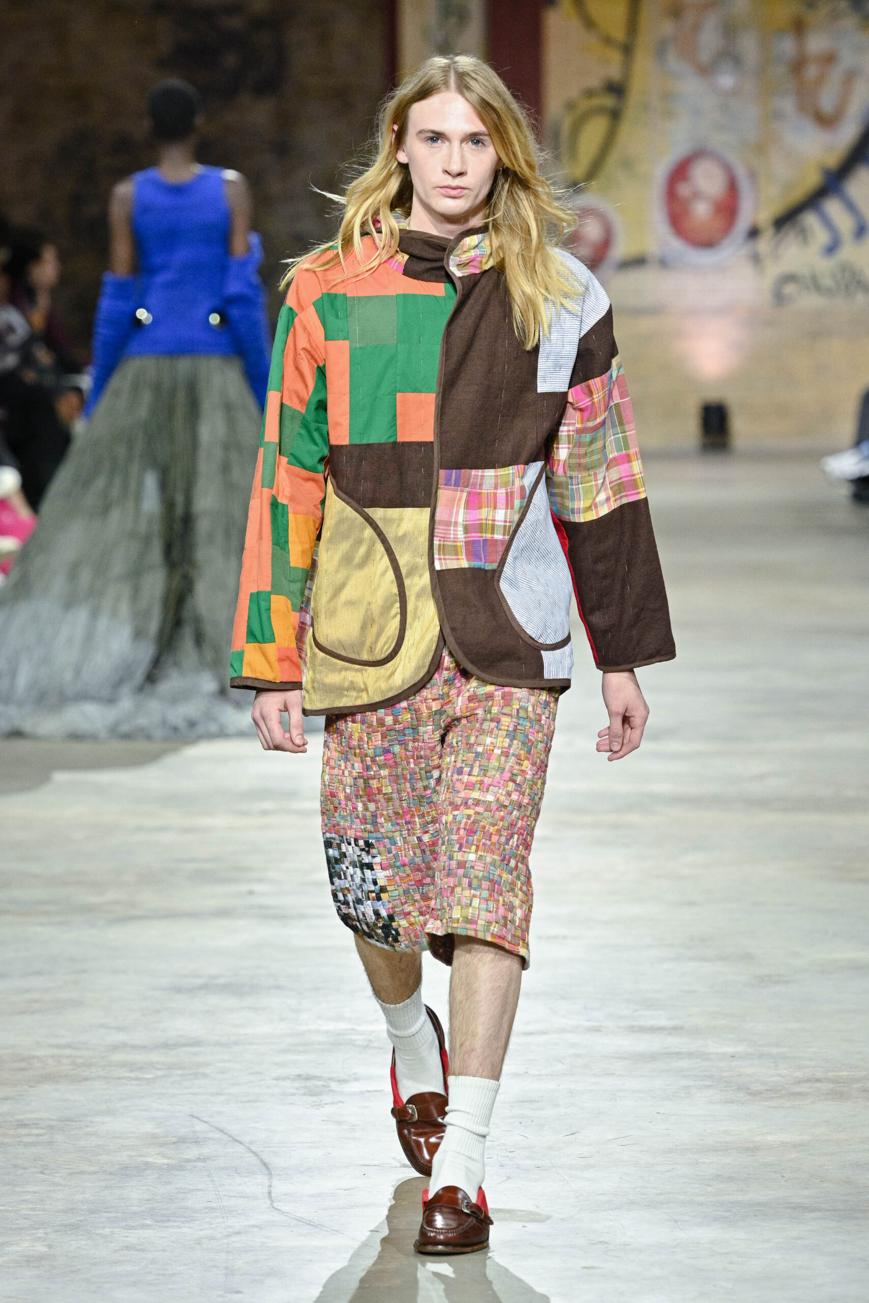 A model walks down a runway wearing an oversized, colorful patchwork jacket in green, brown, orange, and plaid patterns, paired with textured, multicolored shorts. The model completes the look with brown shoes and white socks. In the background, blurred figures and a decorated wall can be seen, indicating a fashion show environment.