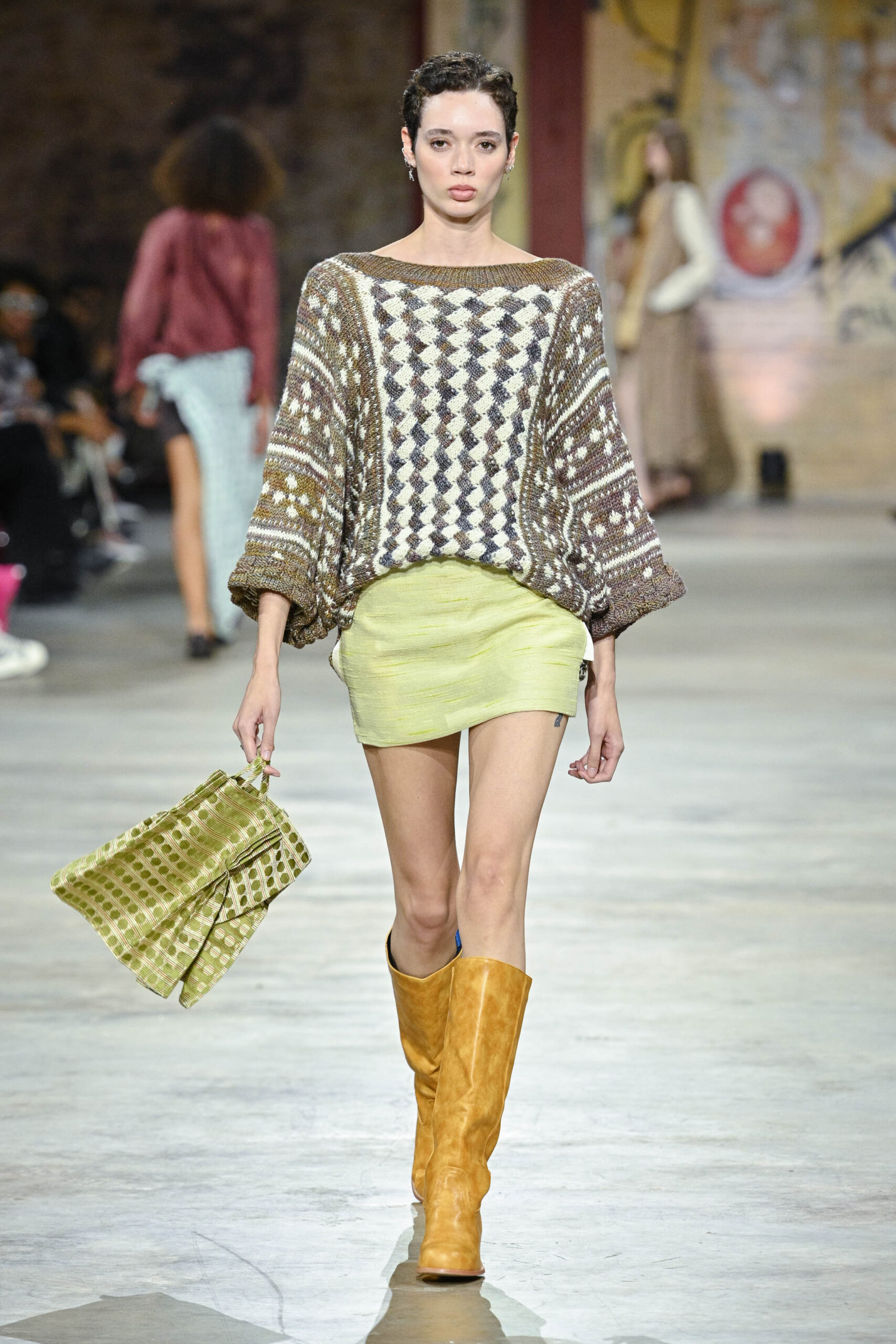 A model walks down the runway wearing a oversized, patterned sweater with a wide neckline, paired with a short, light green skirt. She carries a green patterned handbag and wears knee-high tan leather boots. The runway scene includes blurred figures of other models in the background, with an artistic backdrop and audience seated nearby.