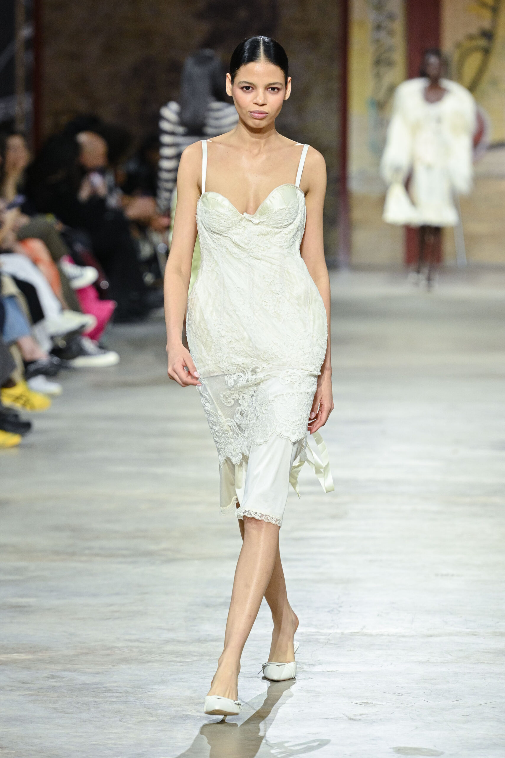 A model walks down a runway wearing a delicate white dress with lace detailing and thin straps. The dress is layered over a light slip, and she pairs it with simple white flats. The background features an audience and another model wearing a different outfit. The setting appears to be a fashion show with a casual, artistic vibe.