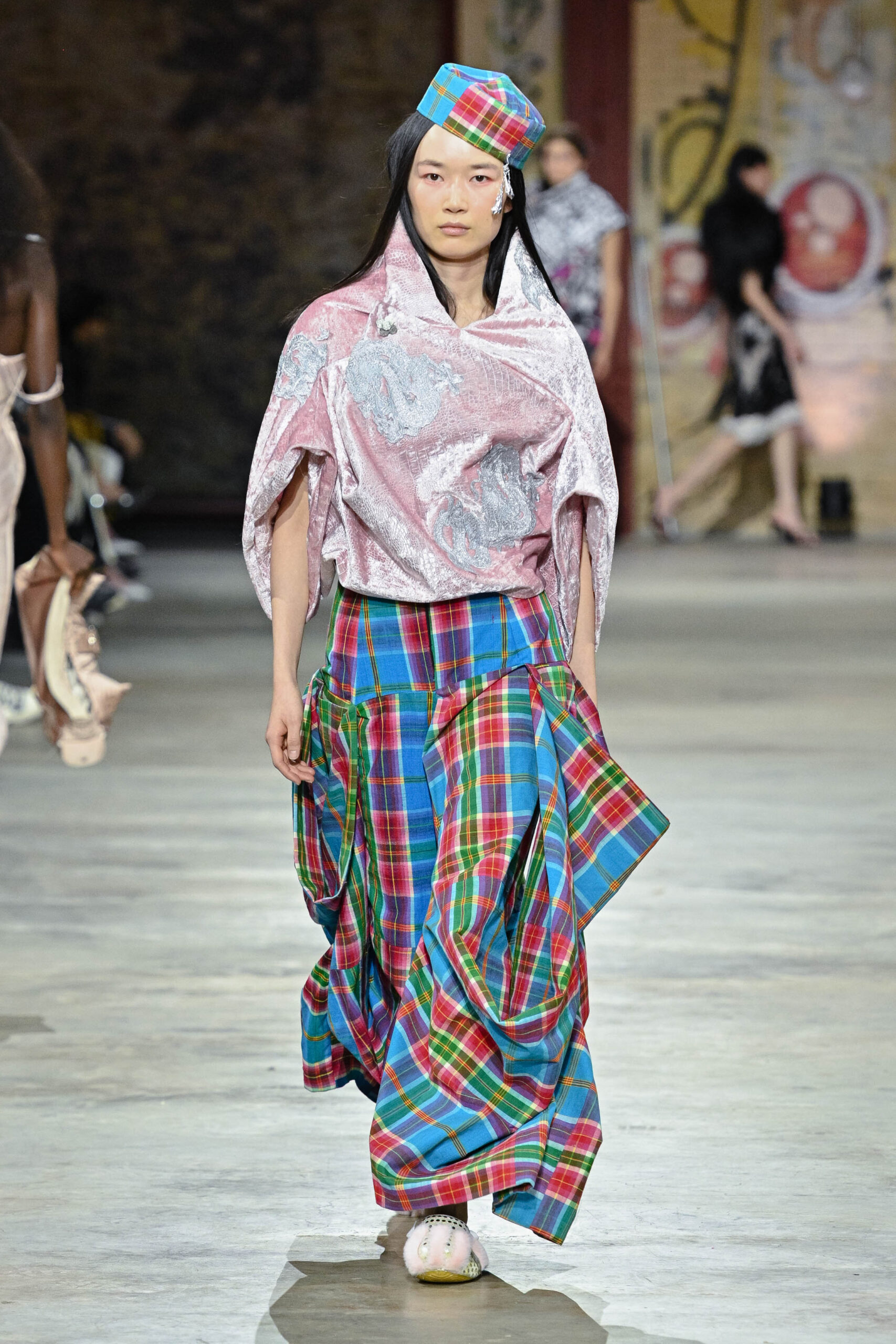 A model walks down the runway wearing a unique outfit composed of a shiny pink top with a loose silhouette and a plaid wrap skirt featuring vibrant colors like blue, red, green, and yellow. The model also sports a colorful plaid hat and fluffy footwear, showcasing an avant-garde fashion style in a spacious, industrial setting with soft lighting.