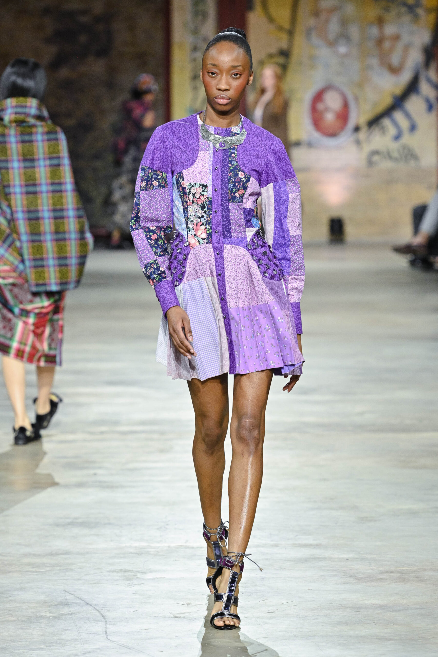 A model walks down a runway, showcasing a vibrant, patchwork dress in shades of purple adorned with floral patterns. The dress features a fitted bodice with long sleeves and a flared skirt. The model wears strappy high-heeled shoes. In the background, other models are seen wearing various patterned outfits in a visually dynamic setting.