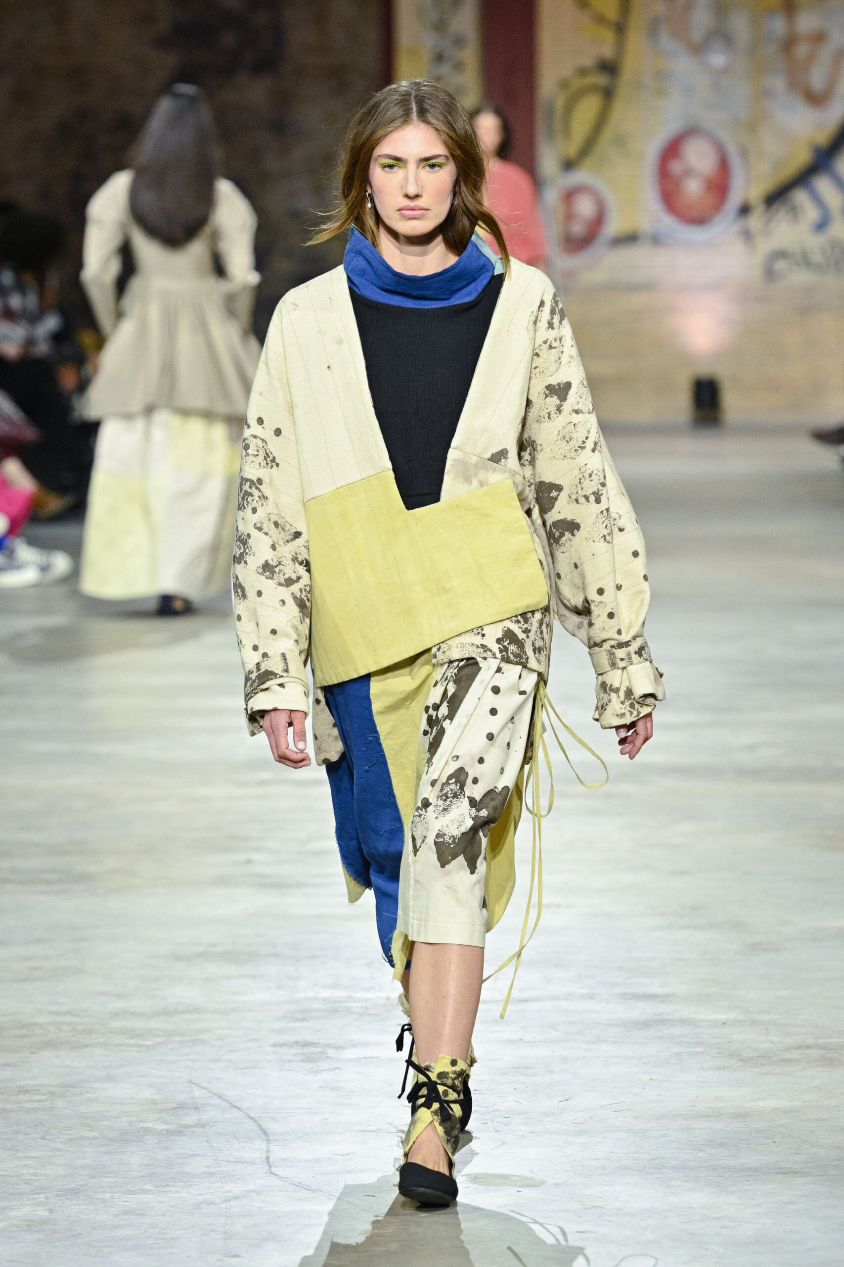 A model walks down a runway wearing a unique, oversized outfit composed of various colors including yellow, blue, and cream. The garment features distinct patterns and textures, highlighting an artistic design. The model has long hair and sports subtle makeup with bright accents. In the background, other models can be seen, with an urban setting characterized by abstract wall art. The runway is well-lit, enhancing the visual impact of the clothing.