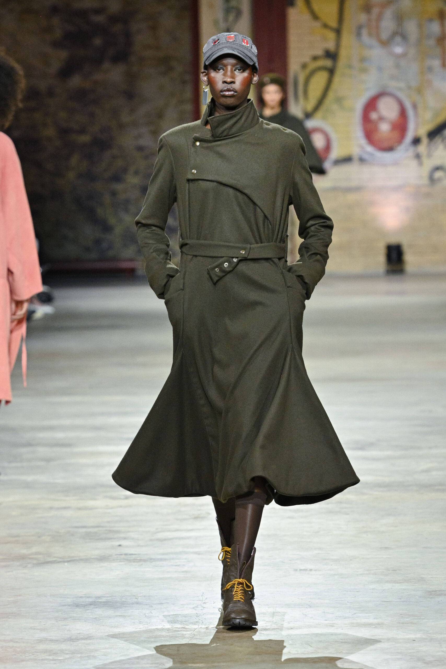 A model walks down a runway in a stylish olive-green trench coat that flares out at the bottom. The coat features a high collar and is cinched at the waist with a belt. The model pairs the outfit with brown lace-up boots with yellow laces and a gray baseball cap. The background shows a faint mural, adding to the fashion show atmosphere.