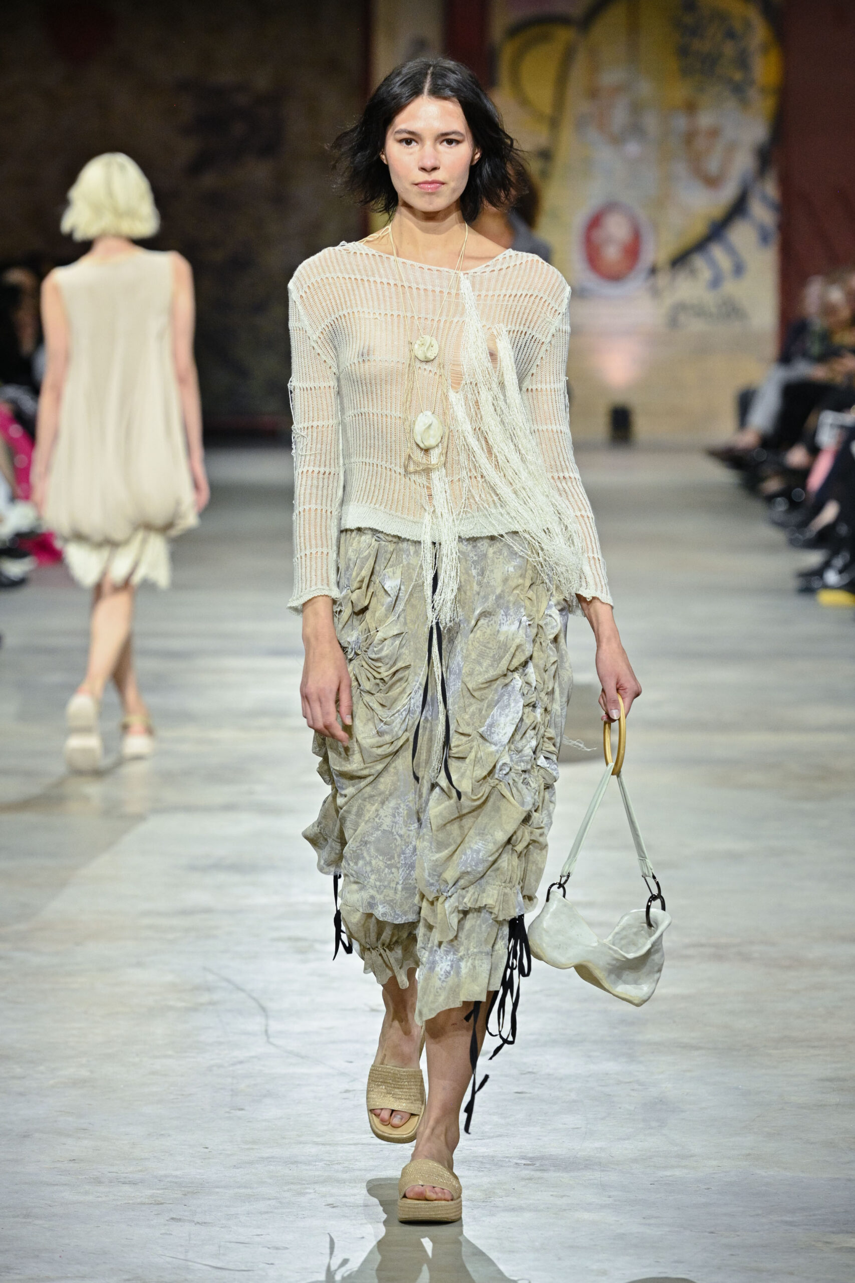 A model walks down a runway wearing a sheer, knitted top with long fringe and a low neckline, paired with a flowy, layered skirt featuring ruffled details. She accessorizes with a small, soft handbag and beige platform sandals. In the background, another model in a light dress is visible, and an audience sits alongside the runway. The setting has artistic wall designs.