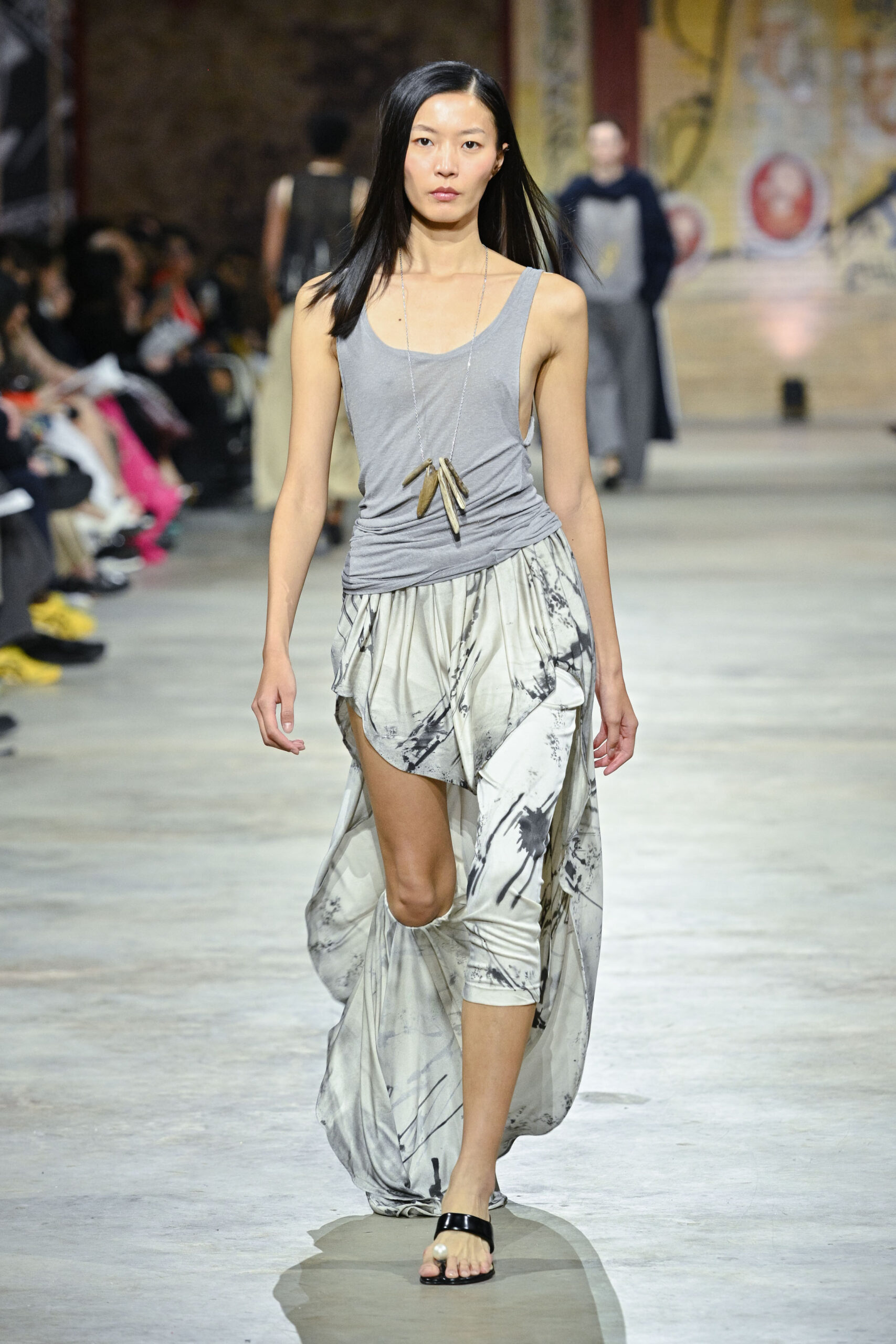A model walks down a runway wearing a gray sleeveless top and a flowing, patterned skirt with layers. The skirt is predominantly light with abstract black designs. She is accessorized with a long necklace featuring metal pendants and is wearing a pair of stylish sandals. The backdrop includes an audience seated around the runway in a spacious venue.