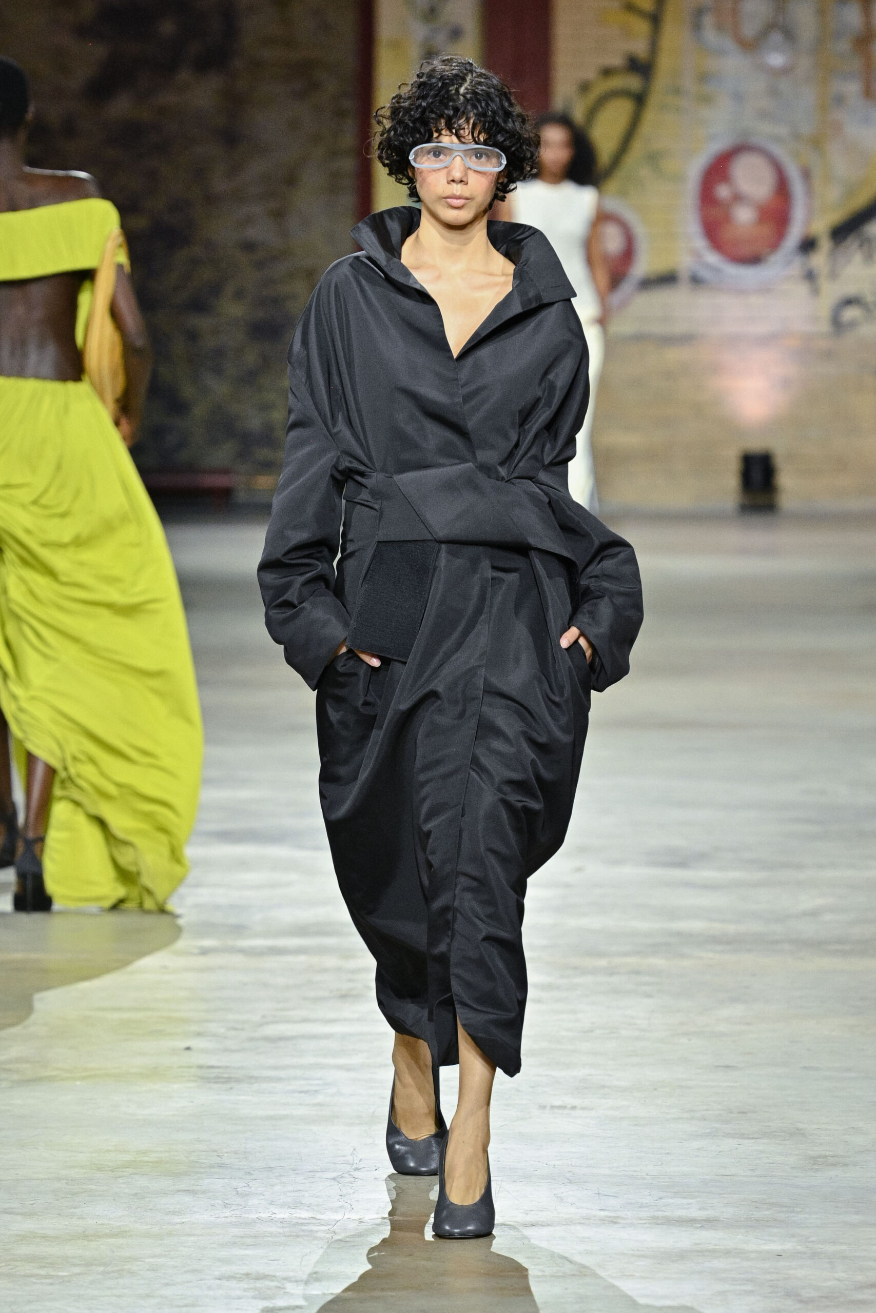 A model walks down the runway wearing a black, oversized dress with a large collar and cinched waist. She has short, curly hair and is accessorized with large, clear glasses. In the background, another model is seen wearing a flowing lime green dress. The runway is set in an industrial space with textured walls.