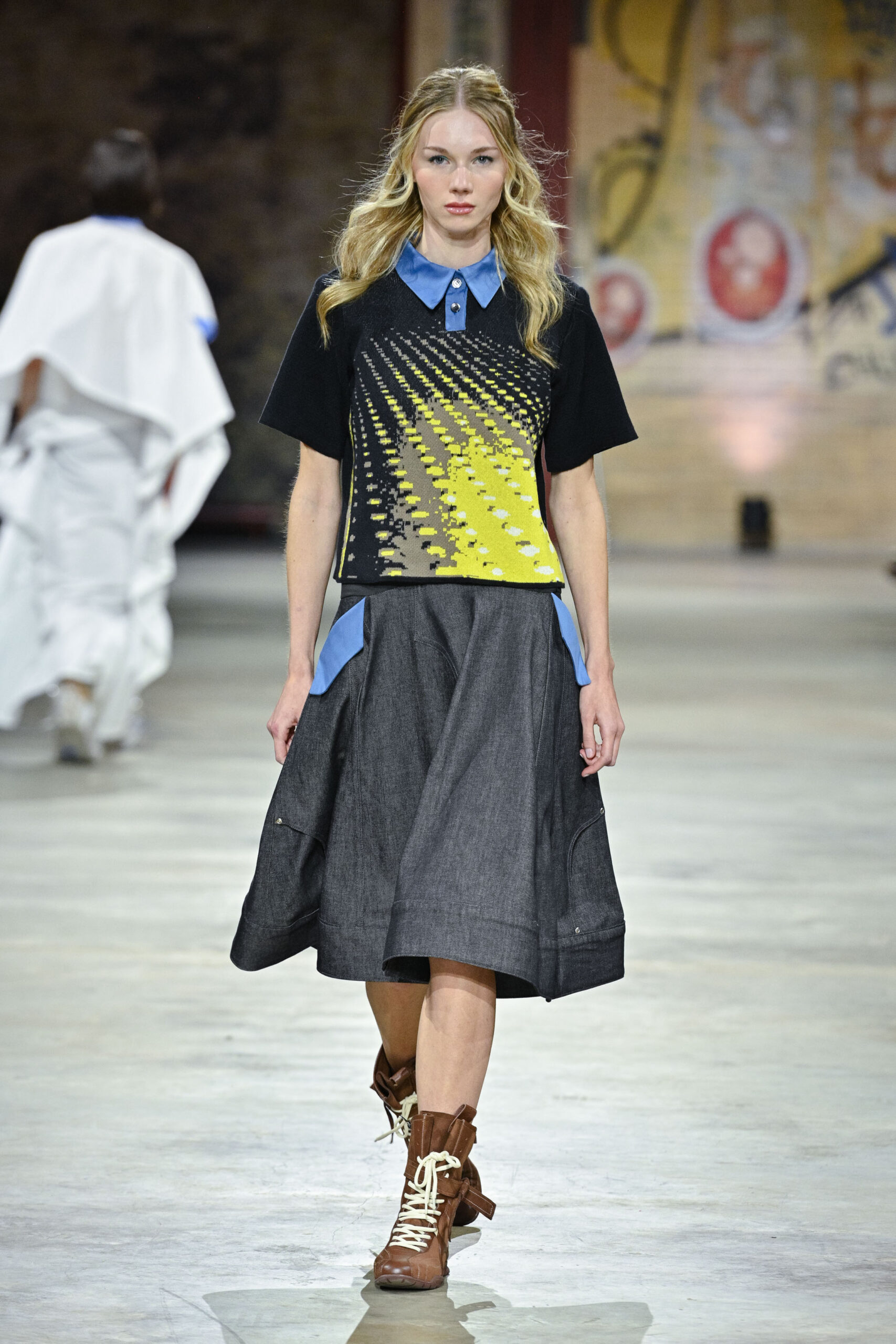A model walks down a runway wearing a stylish outfit that consists of a short-sleeved black top with a yellow and black graphic design and a blue collared neckline, paired with a dark grey skirt featuring blue pockets. She completes the look with brown, knee-high lace-up boots. The background features a blurred figure in white and colorful wall art.