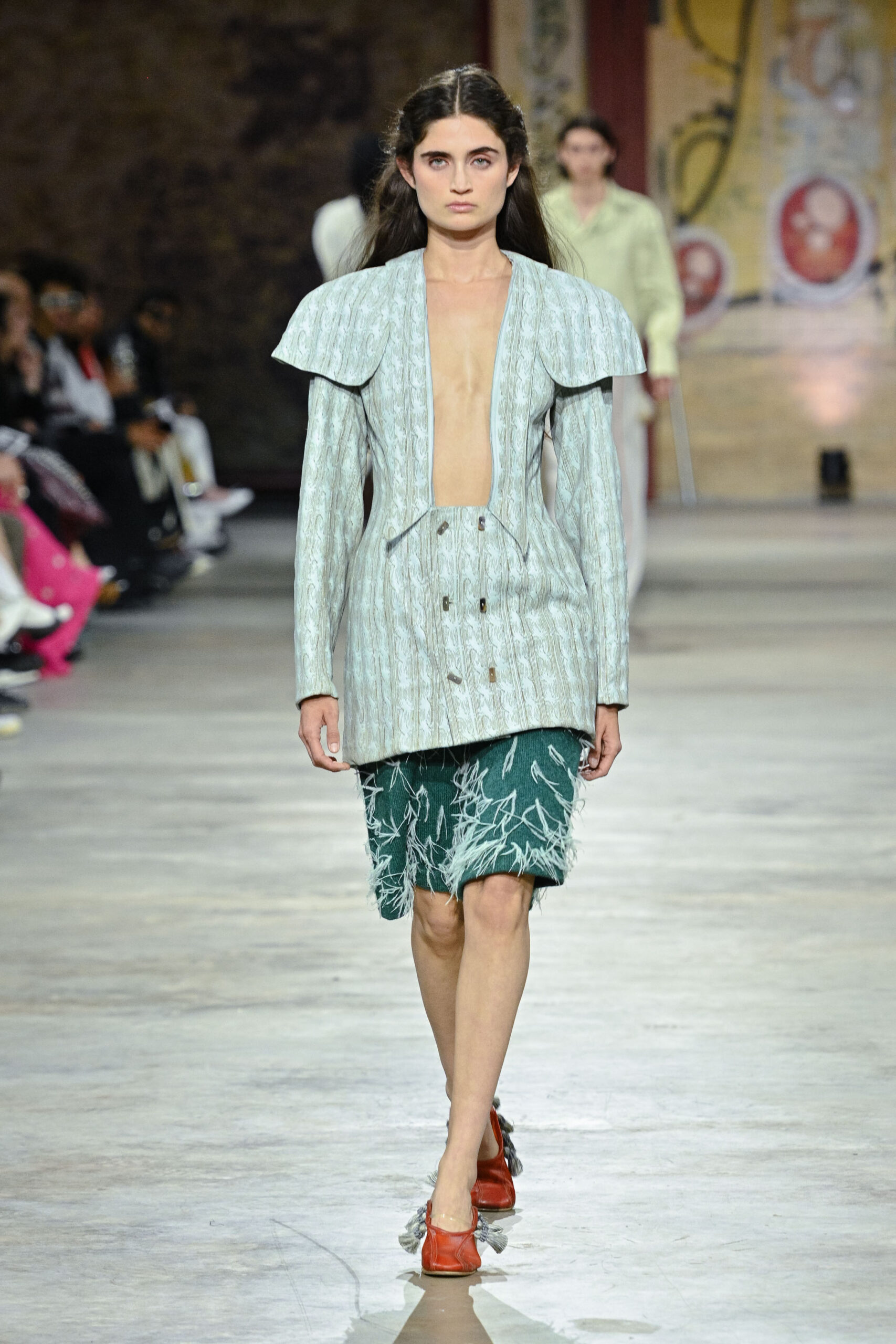 A model walks down a runway wearing a light blue oversized jacket with a deep neckline and matching patterned green skirt with frayed edges. She pairs the outfit with red shoes that have tassels. In the background, an audience is seated, with other models visible in the distance. The setting has a rustic, artistic vibe.