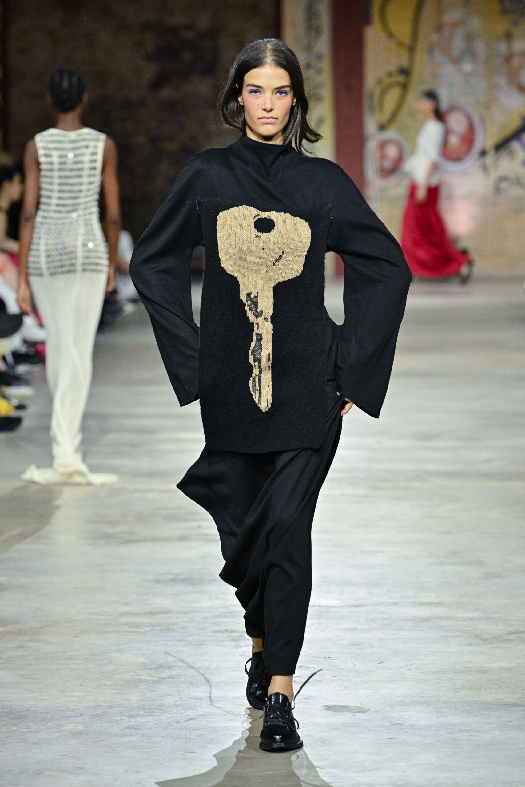 A model walks down a runway wearing a black oversized sweater featuring a large key design in beige. The sweater has wide sleeves and is paired with loose black pants and shiny black shoes. In the background, another model can be seen wearing a form-fitting, shimmering outfit, while additional models are present further back, showcasing various designs. The setting appears to be an industrial space with artistic murals.