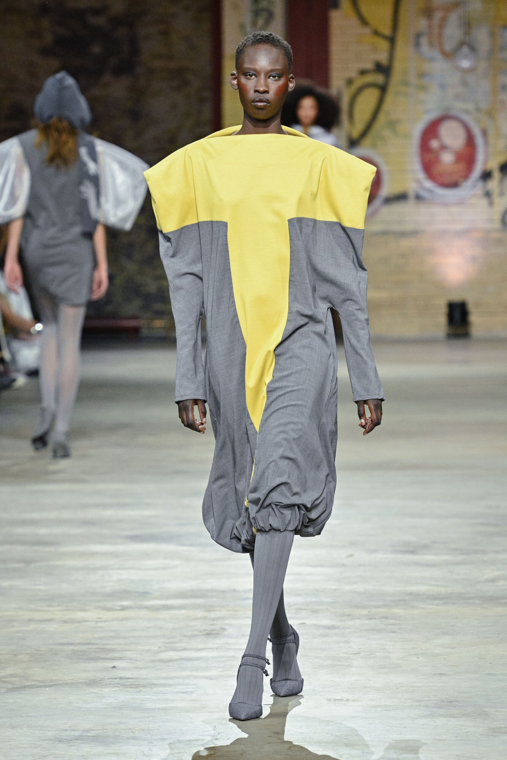 A model walks down a runway, showcasing an avant-garde outfit composed of gray and yellow fabric. The dress features a large yellow collar and a boxy silhouette that extends to the knees, with voluminous sleeves and a gathered hem. The model pairs the dress with matching gray thigh-high socks and heels. In the background, another model is partially visible, wearing a gray outfit. The setting appears to be a fashion event with an industrial backdrop.