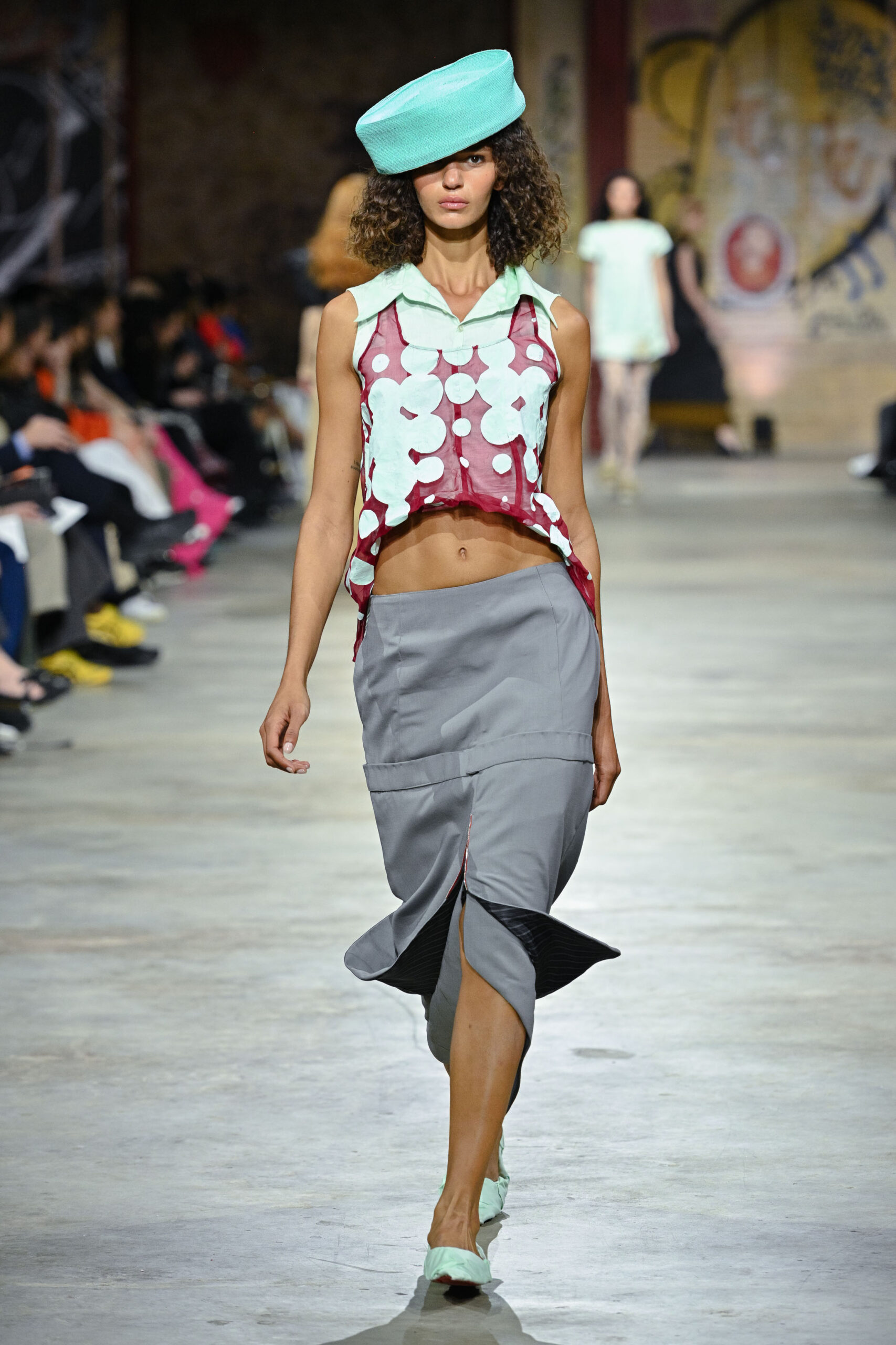 A model walks down a runway wearing a unique outfit composed of a cropped, sleeveless top with large white polka dots on a red background and a gray skirt featuring a slit. She accessorizes with a large, mint green hat and matching mint green shoes. The scene is set in a fashion show environment with an audience in the background.