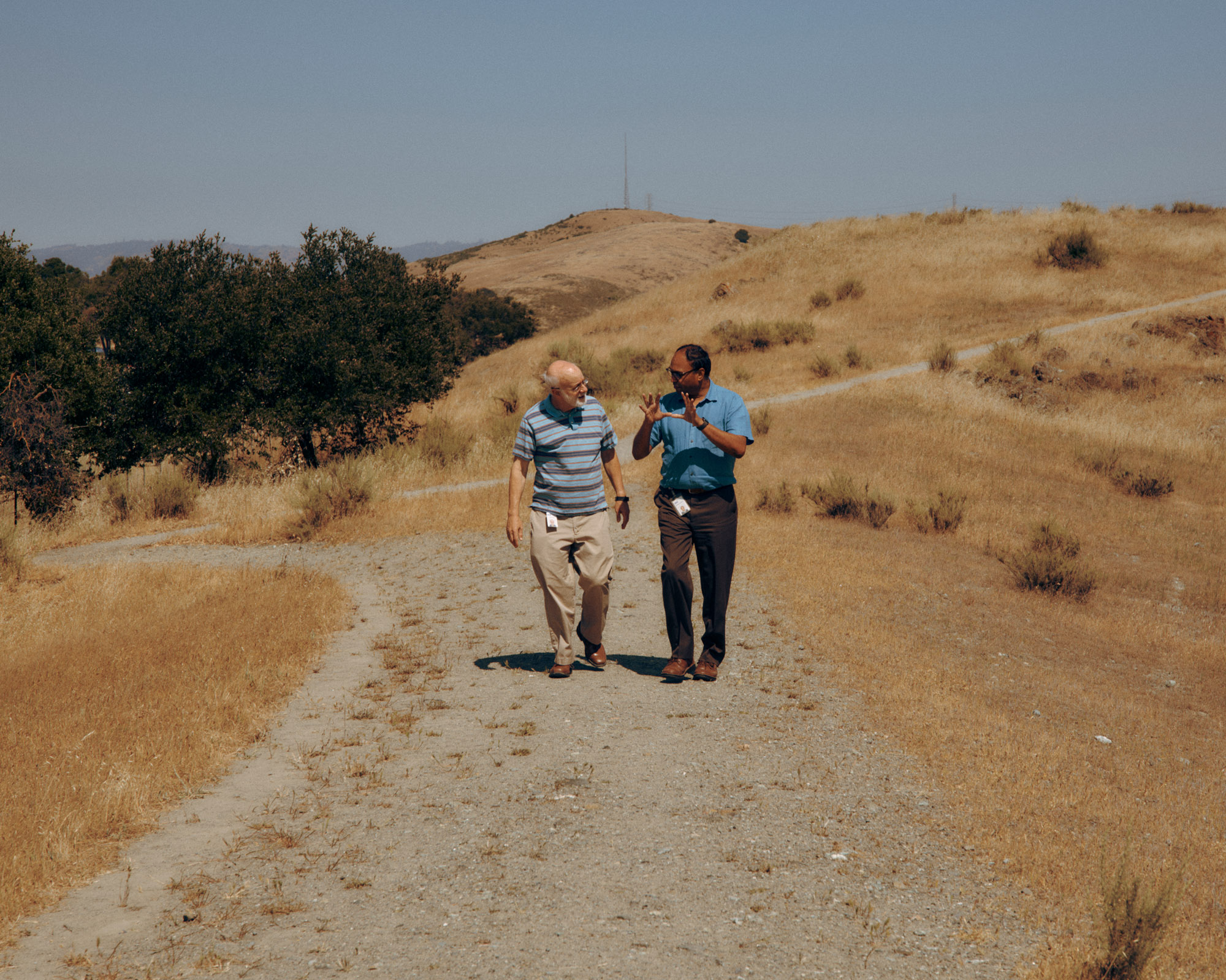 Two people are walking on a gravel path in a dry, hilly landscape. One person, wearing a blue shirt, gestures with their hands as they talks to the other person, who is dressed in a striped shirt and light-colored pants. The background features low hills and sparse vegetation under a clear blue sky.