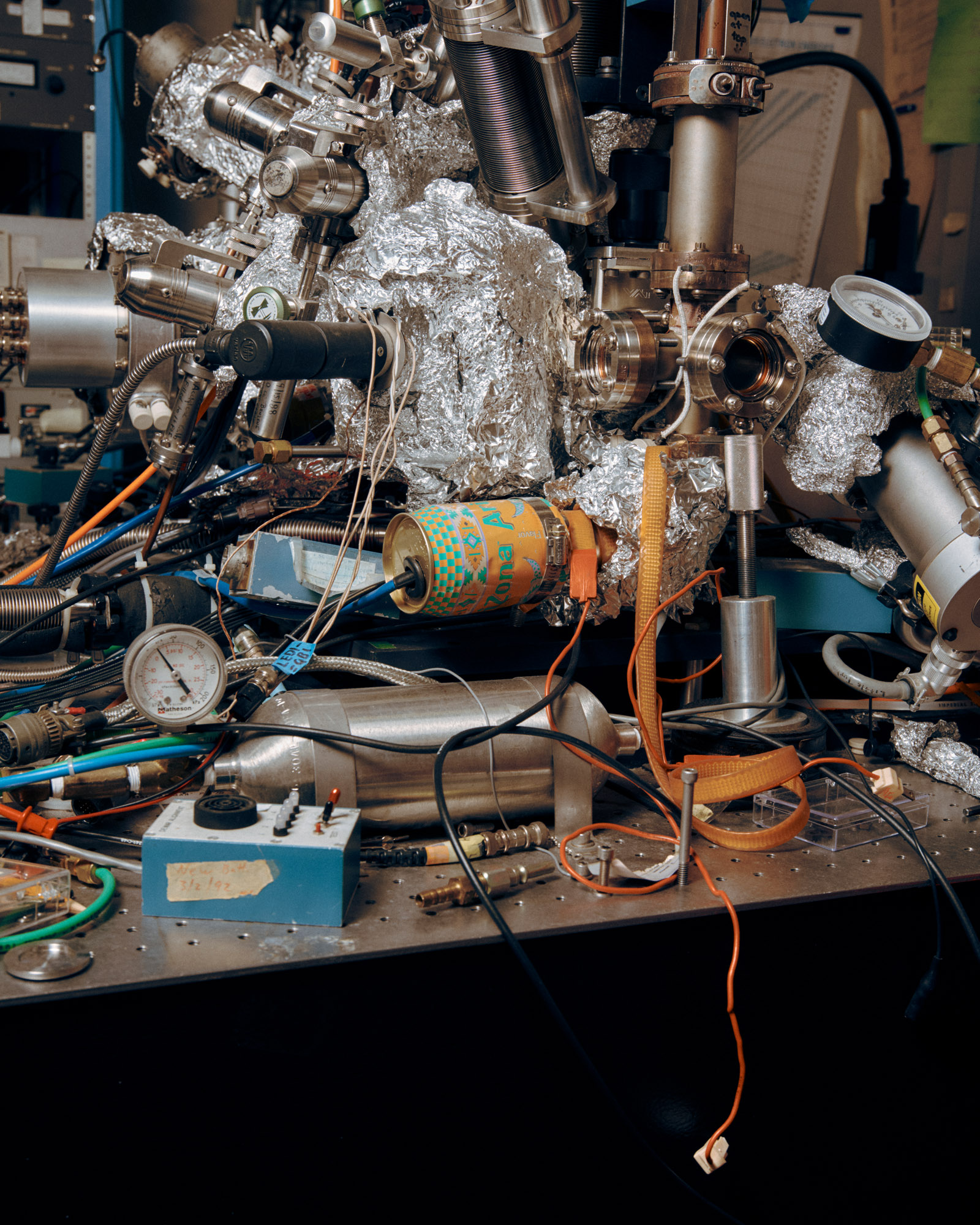 A cluttered laboratory workspace filled with various scientific instruments and equipment. There are metal tubes, valves, and connectors, along with wires and cables crisscrossing the surface. An oxygen tank and a pressure gauge are visible alongside a blue control box with a label. Tinfoil and colorful cans are interspersed among the equipment.
