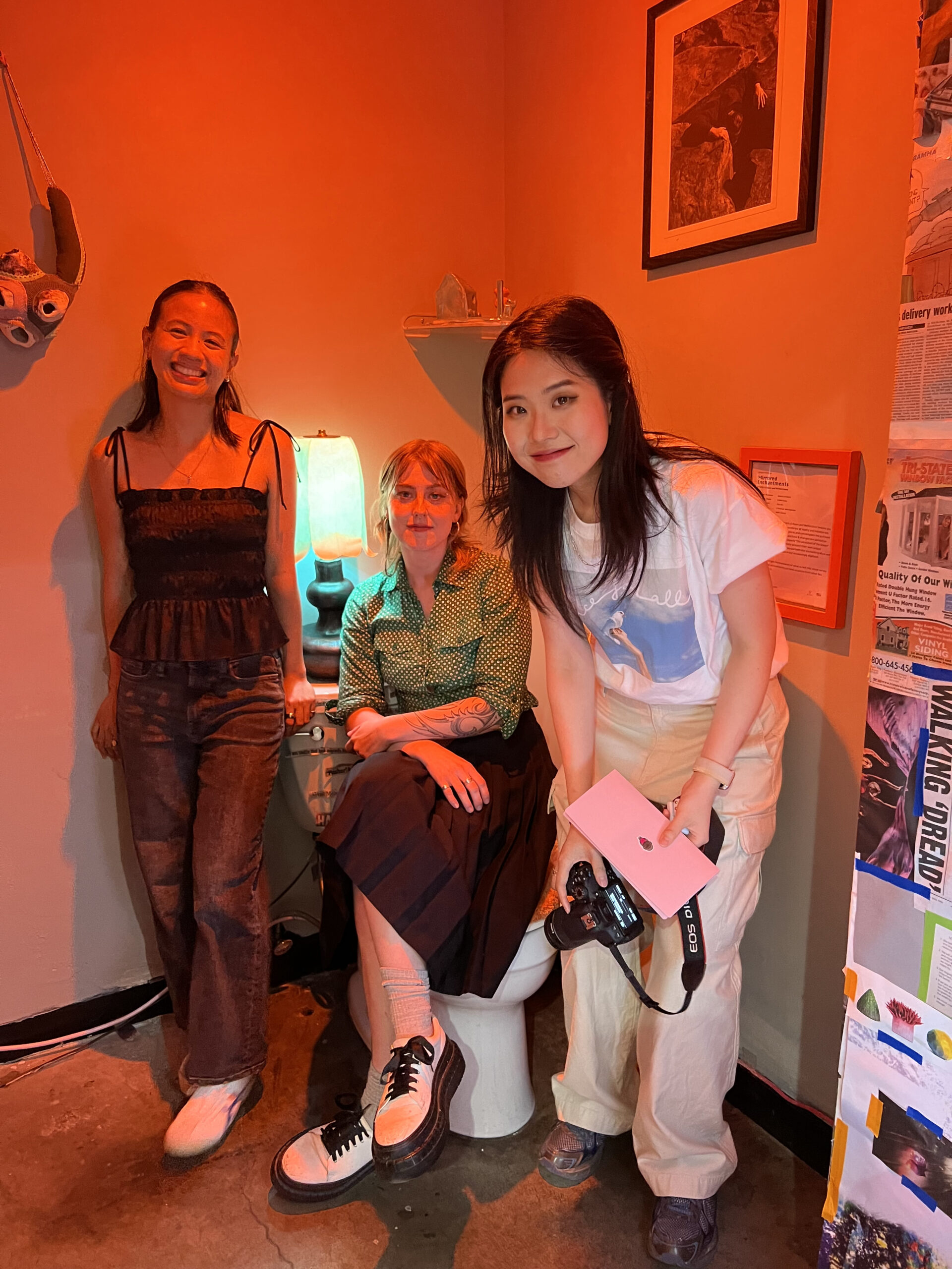 Three people pose together in a red-lit room. One stands on the left, smiling, dressed in a black top and dark jeans. The person in the center sits on a toilet, wearing glasses and a green polka dot shirt with a black skirt. The third stands on the right, holding a camera and a pink notebook, wearing a white T-shirt and light pants. A colorful collage is visible in the background.