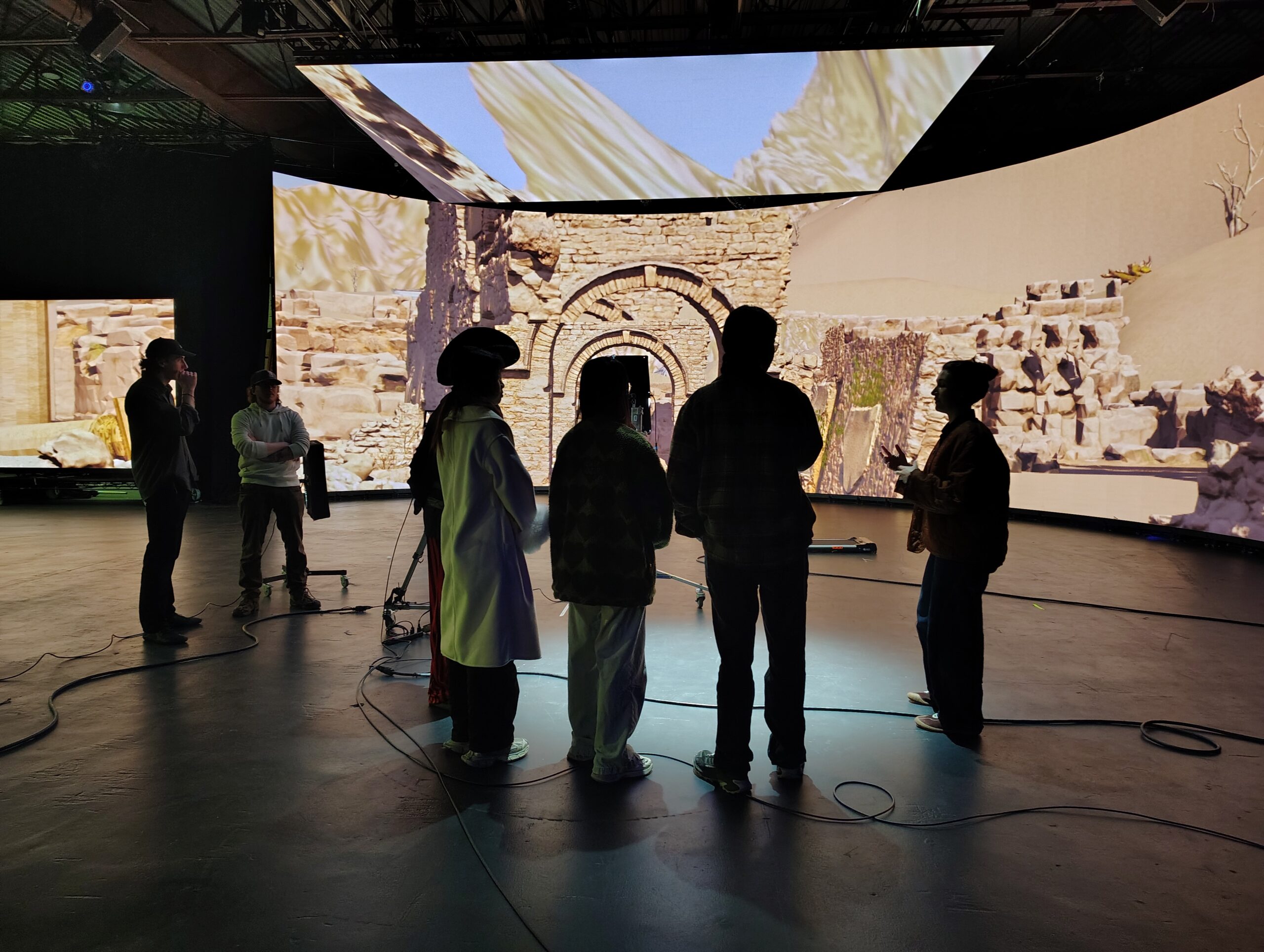 A group of five people stands in front of a large, curved digital screen displaying a virtual landscape. The scene shows ancient ruins and desert-like terrain. The individuals are engaged in discussion, with various equipment visible in the foreground, including cables and a camera on a tripod.
