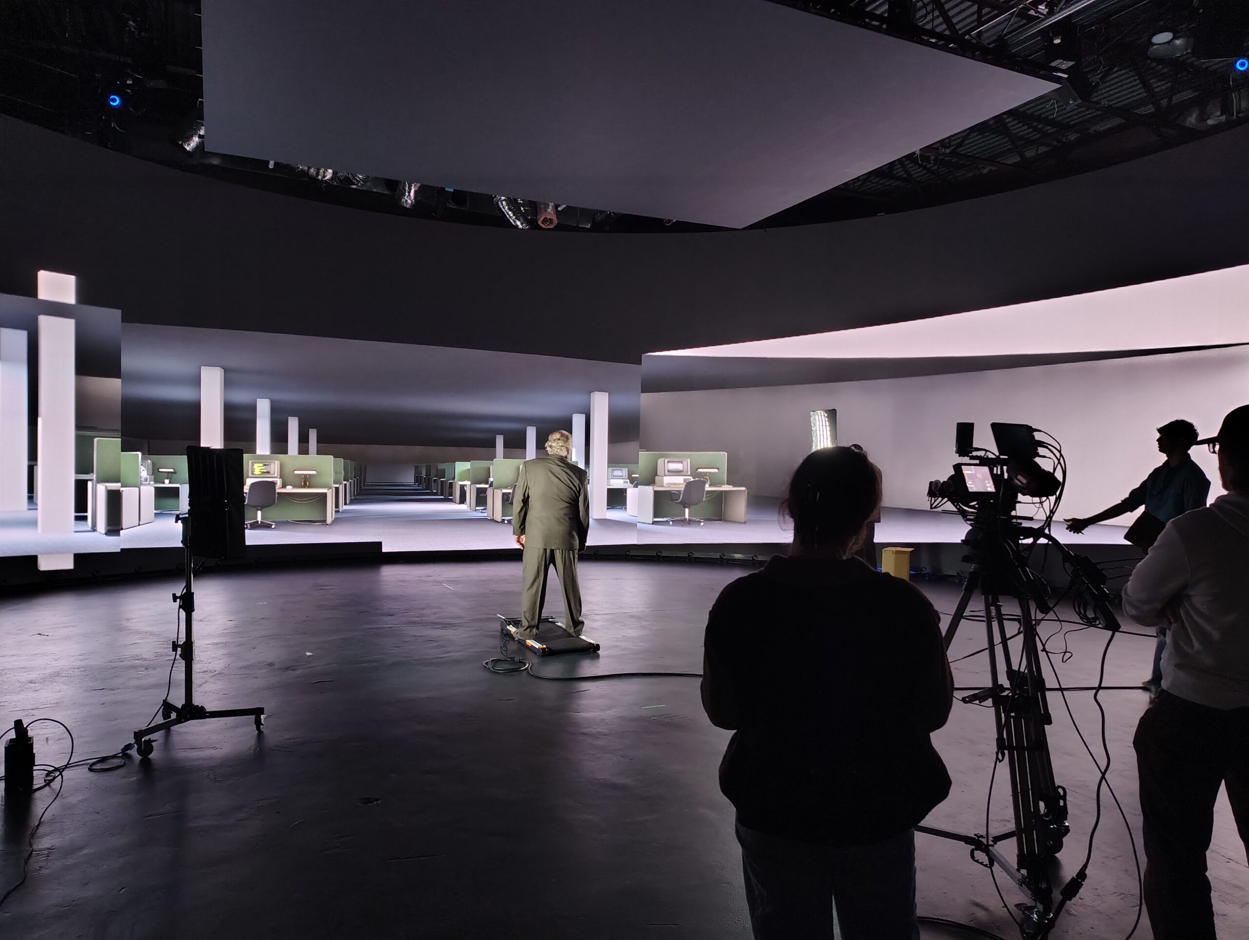 A professional studio setting with a man in a suit standing on a platform, facing a large digital backdrop displaying an office layout with desks and chairs. In the foreground are three individuals: one operating a camera, and two others observing, with equipment visible in the background. The environment is dimly lit, emphasizing the illuminated screens.