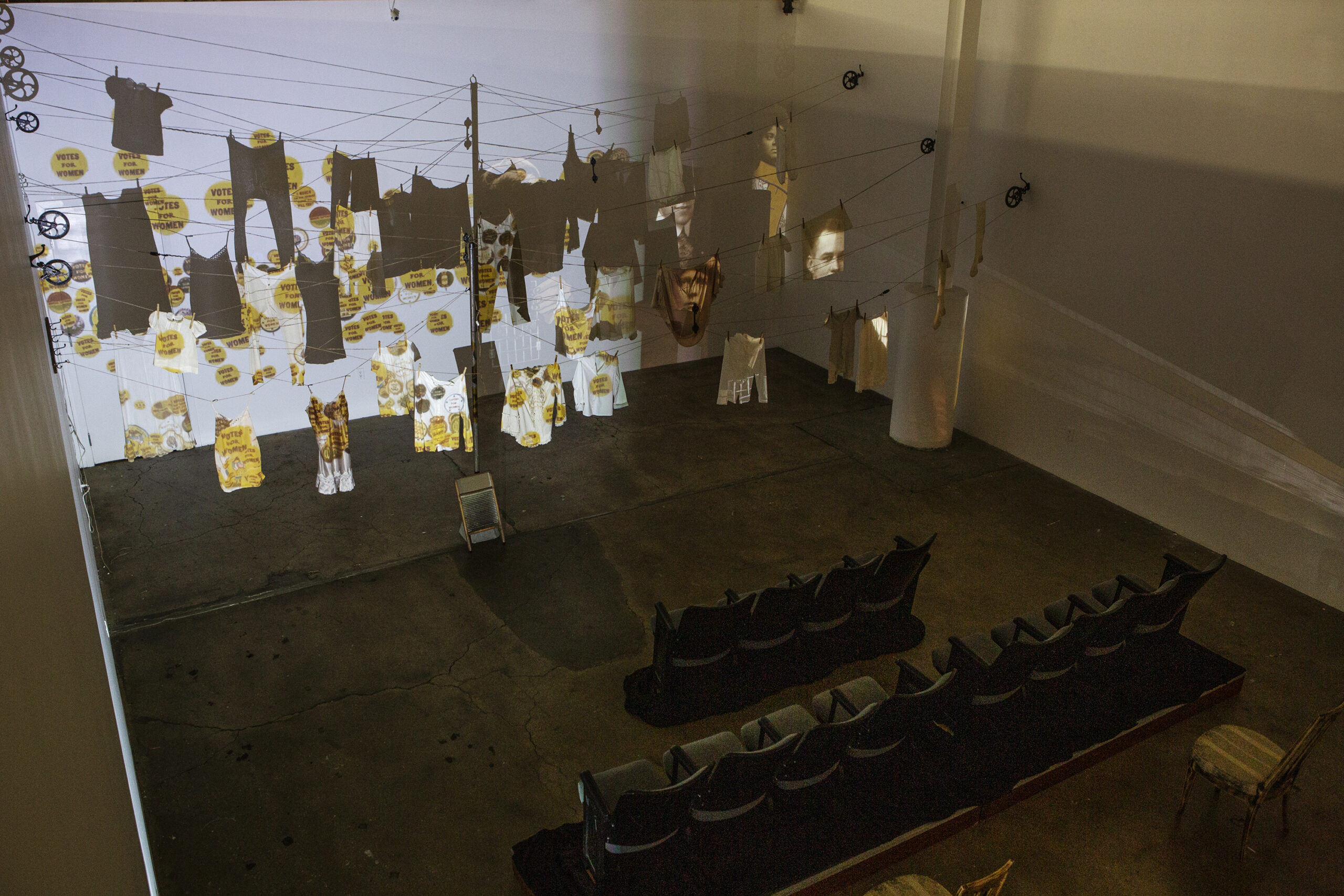 An art installation features clothes suspended on wires, with projections of images and text on them. The space includes empty seats arranged in rows in front of the installation.